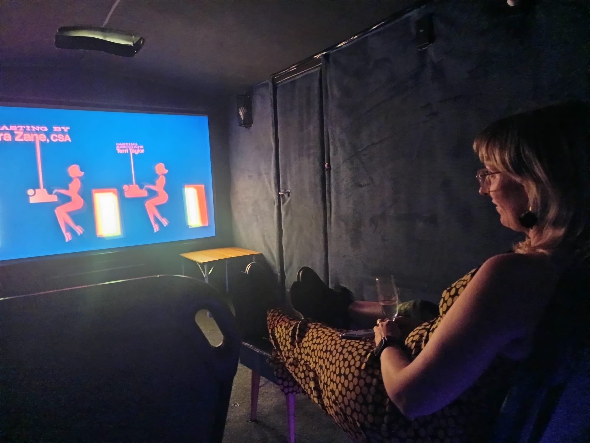 A woman sitting in Mini Cine caravan watching a movie, with a large blue screen displaying pink and white graphics and text.