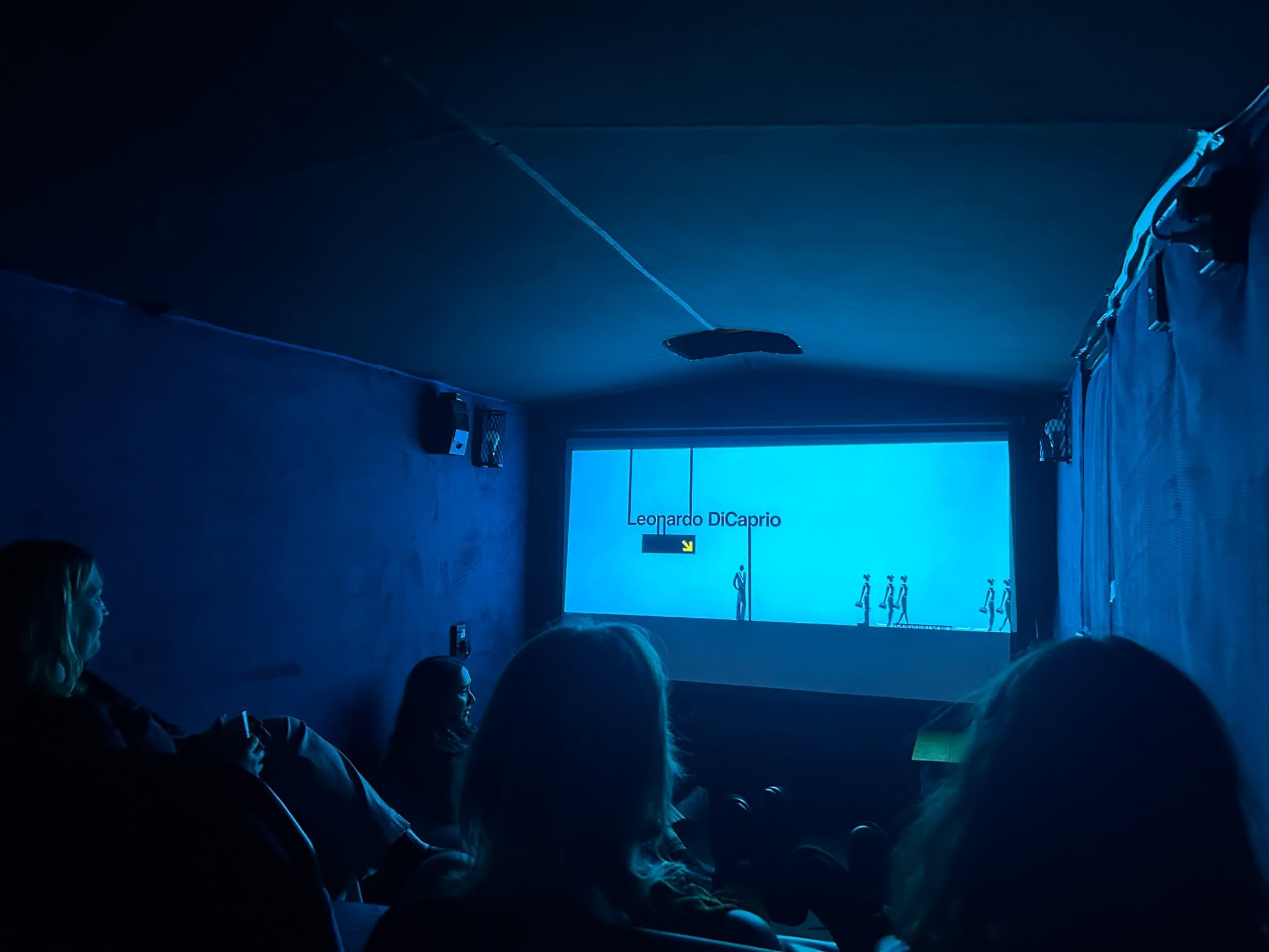 Darkened room with people seated, large screen displaying 'Leonardo DiCaprio' at the front, and blue lighting.