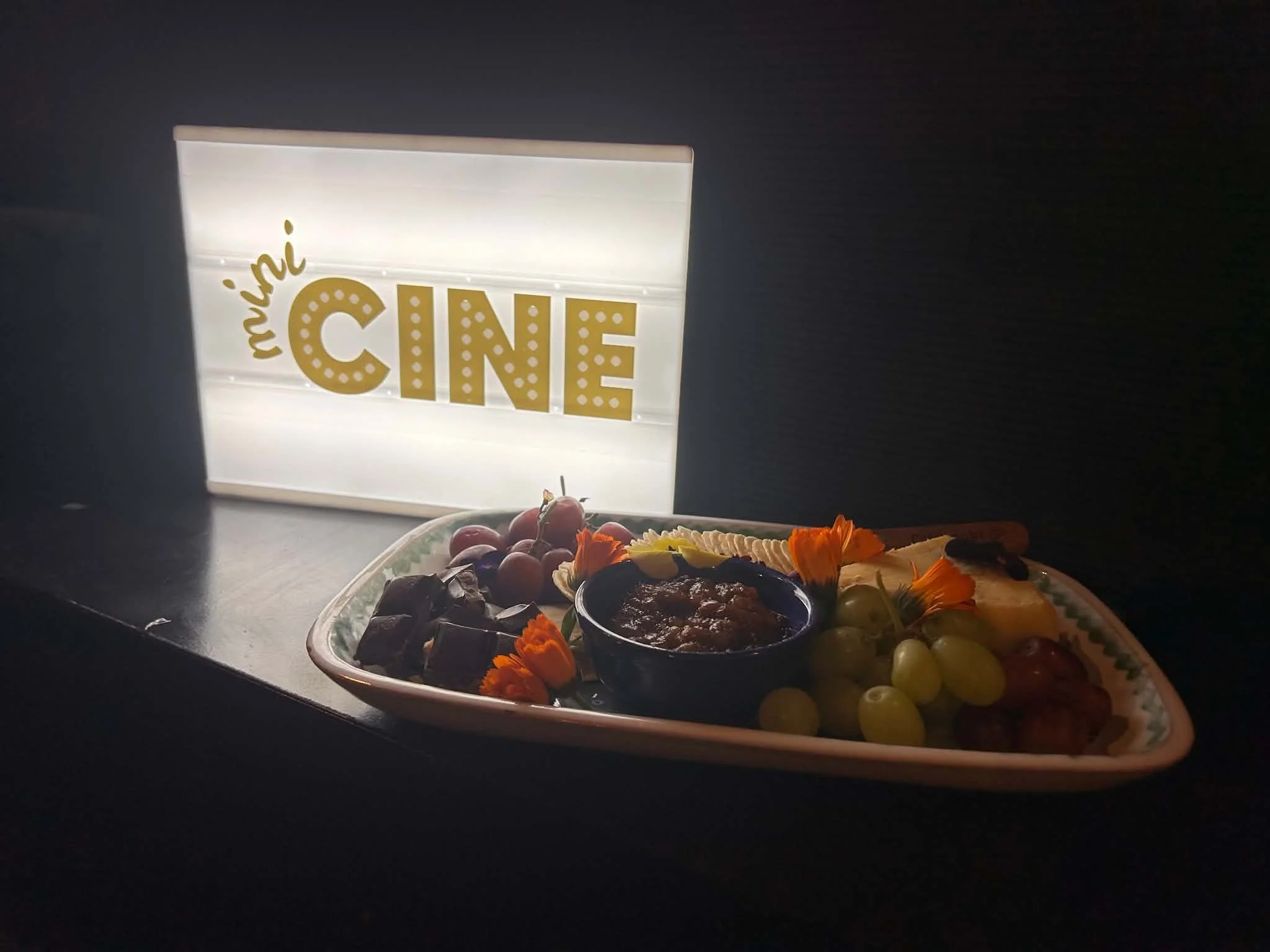 A lively indoor scene of a movie theater with a backlit sign reading 'MINI CINE', and snack tray in the foreground.