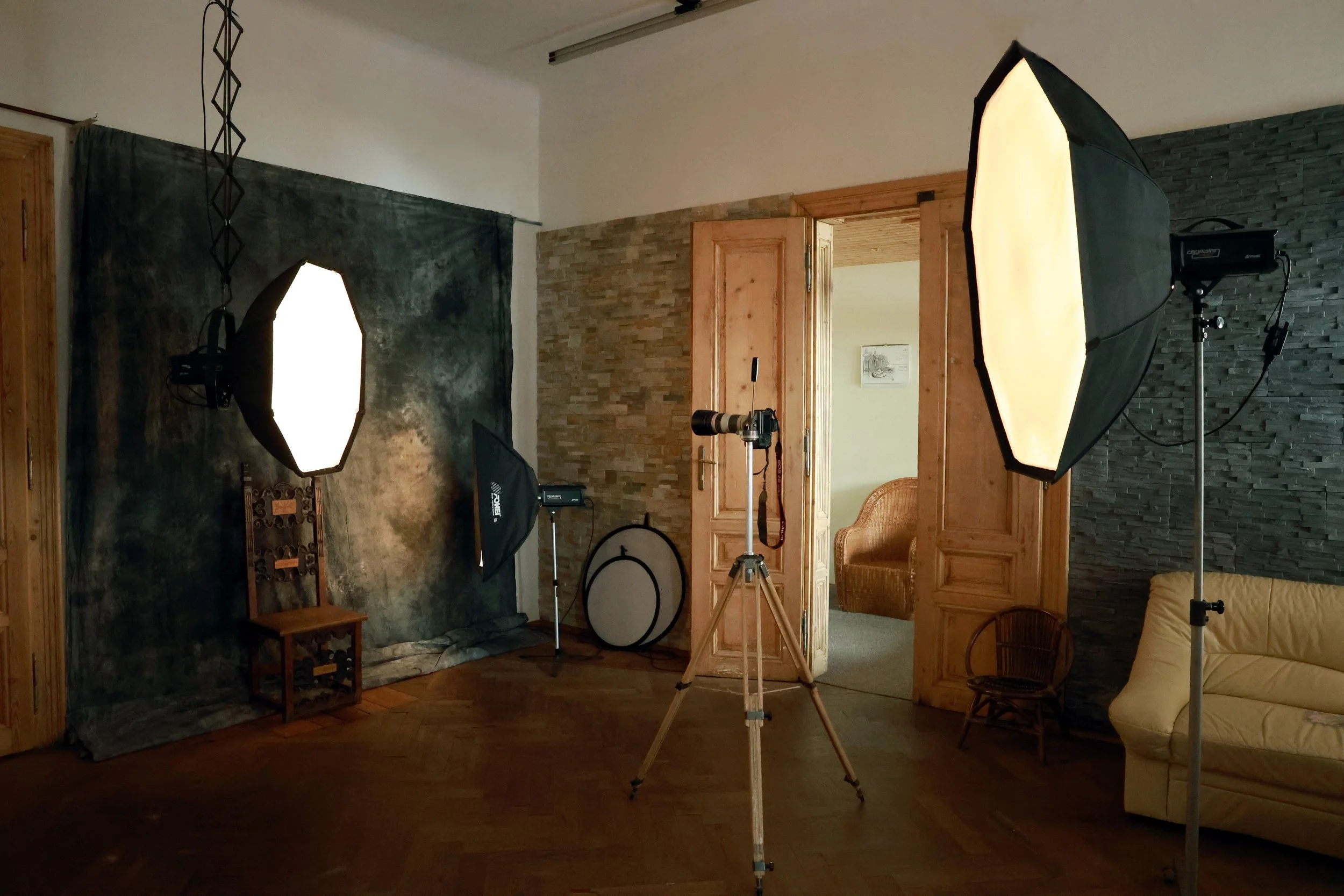 A photography setup in a room with studio lights, a camera on a tripod, a backdrop, and furniture like a chair and a sofa, indicating a photo or video shoot environment.