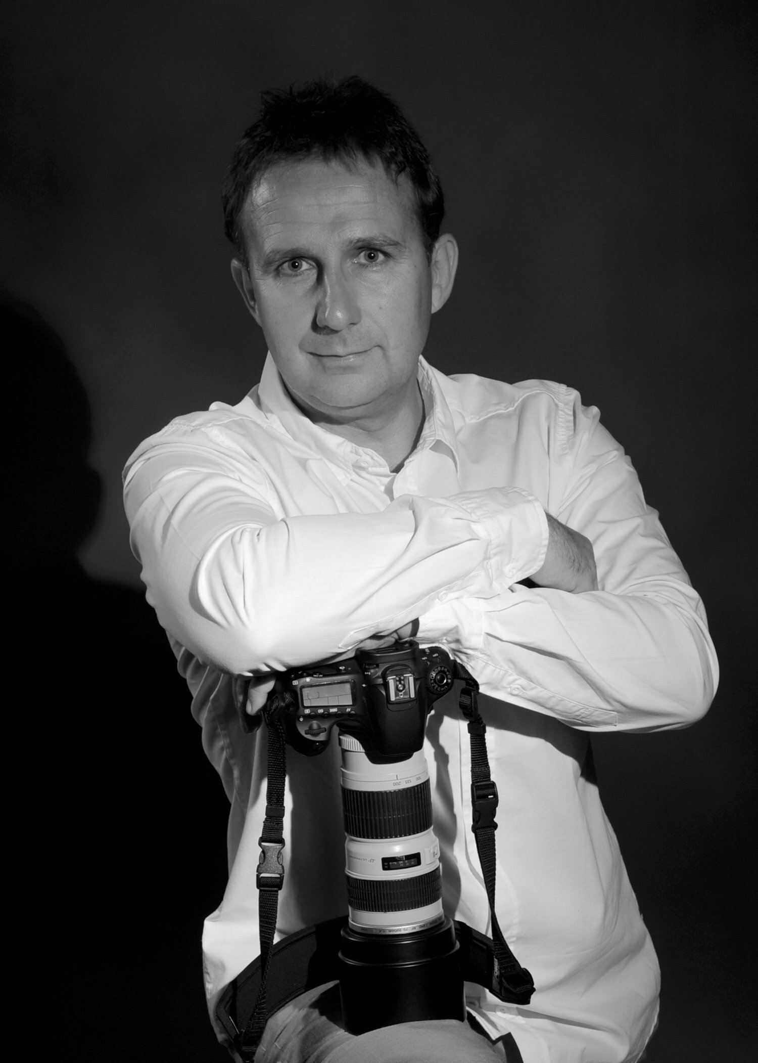 Black and white photo of a man with short dark hair and light skin, wearing a white shirt, sitting with his arms crossed over a camera with a large telephoto lens, looking directly at the camera with a slight smile, against a dark background.