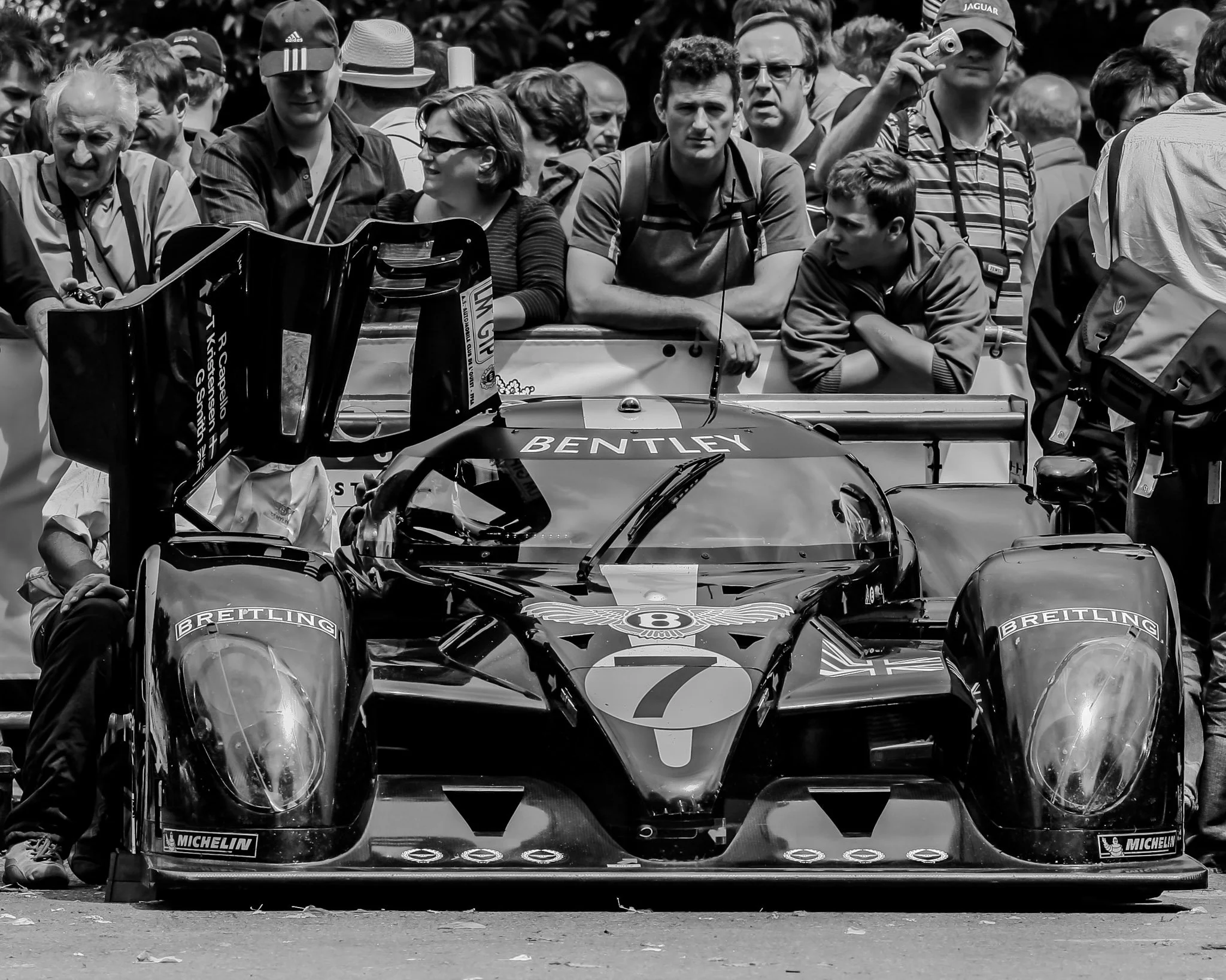 Black and white photo of a crowd of people gathered around a race car at a motorsport event, with some individuals taking photos or videos. The race car has branding including Bentley and Michelin.