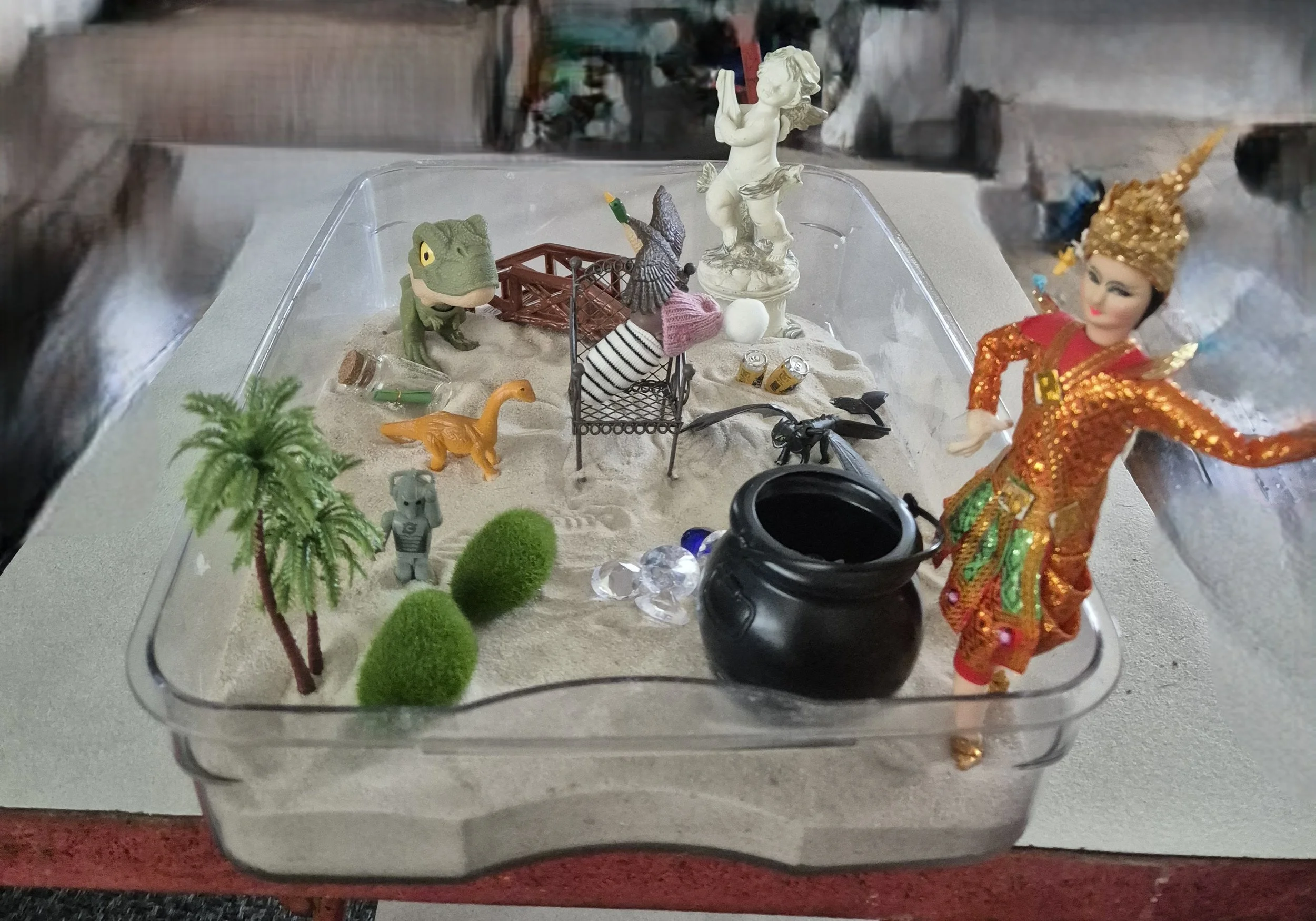 Miniature scene with toy dinosaurs, a cherub statue, small trees, and various objects in a clear plastic container.