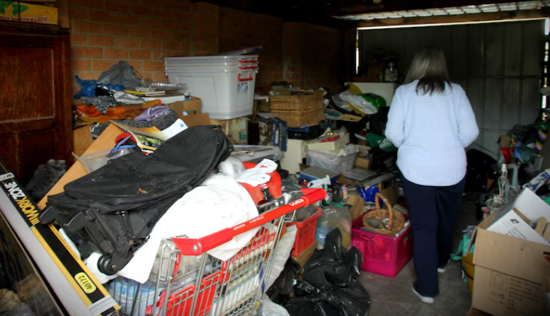 A woman with gray hair wearing a light blue jacket walking through a cluttered garage filled with boxes, bags, and miscellaneous items.