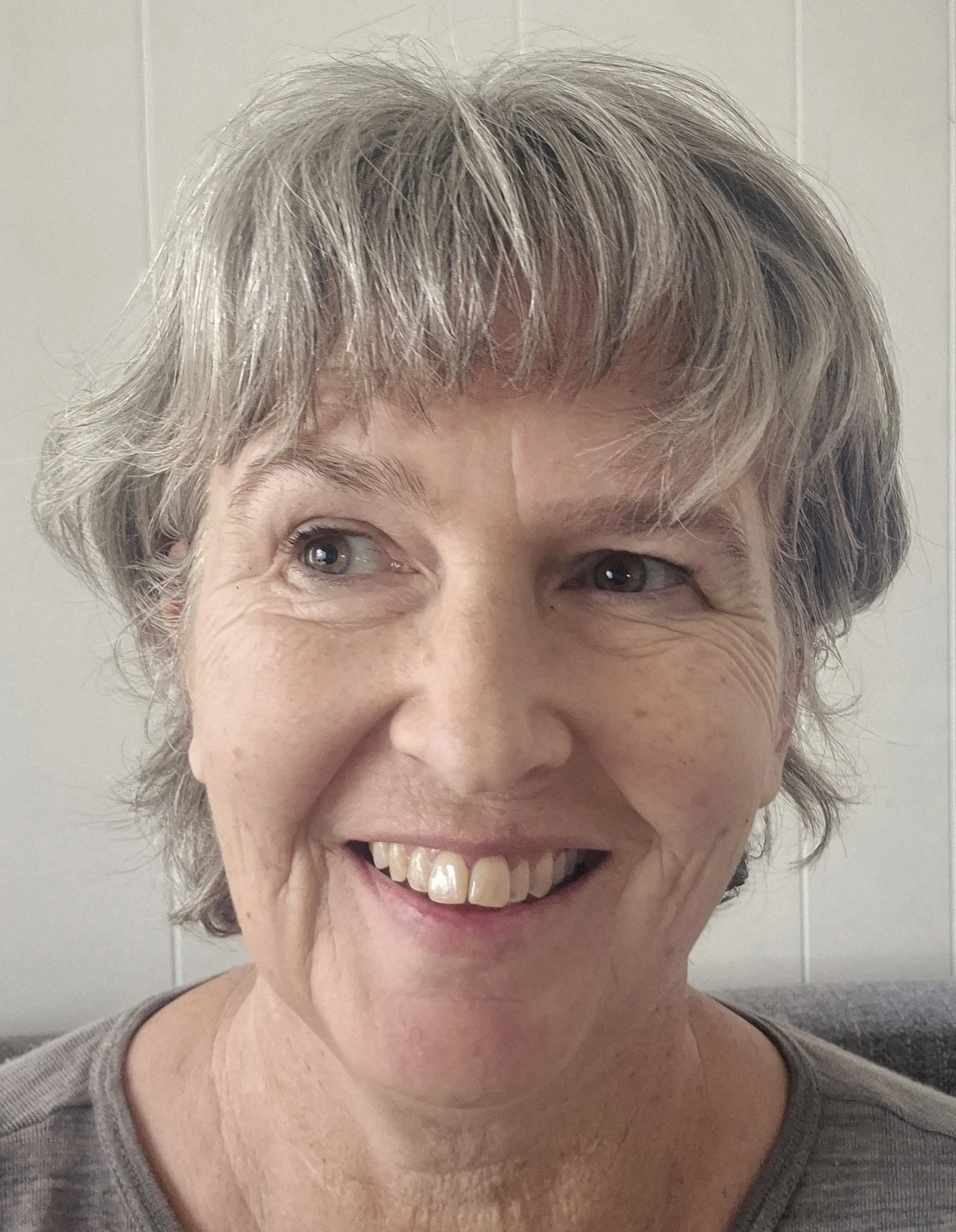 Close-up of a cheerful woman with short, gray and blonde hair, smiling and showing her teeth, wearing a gray top.