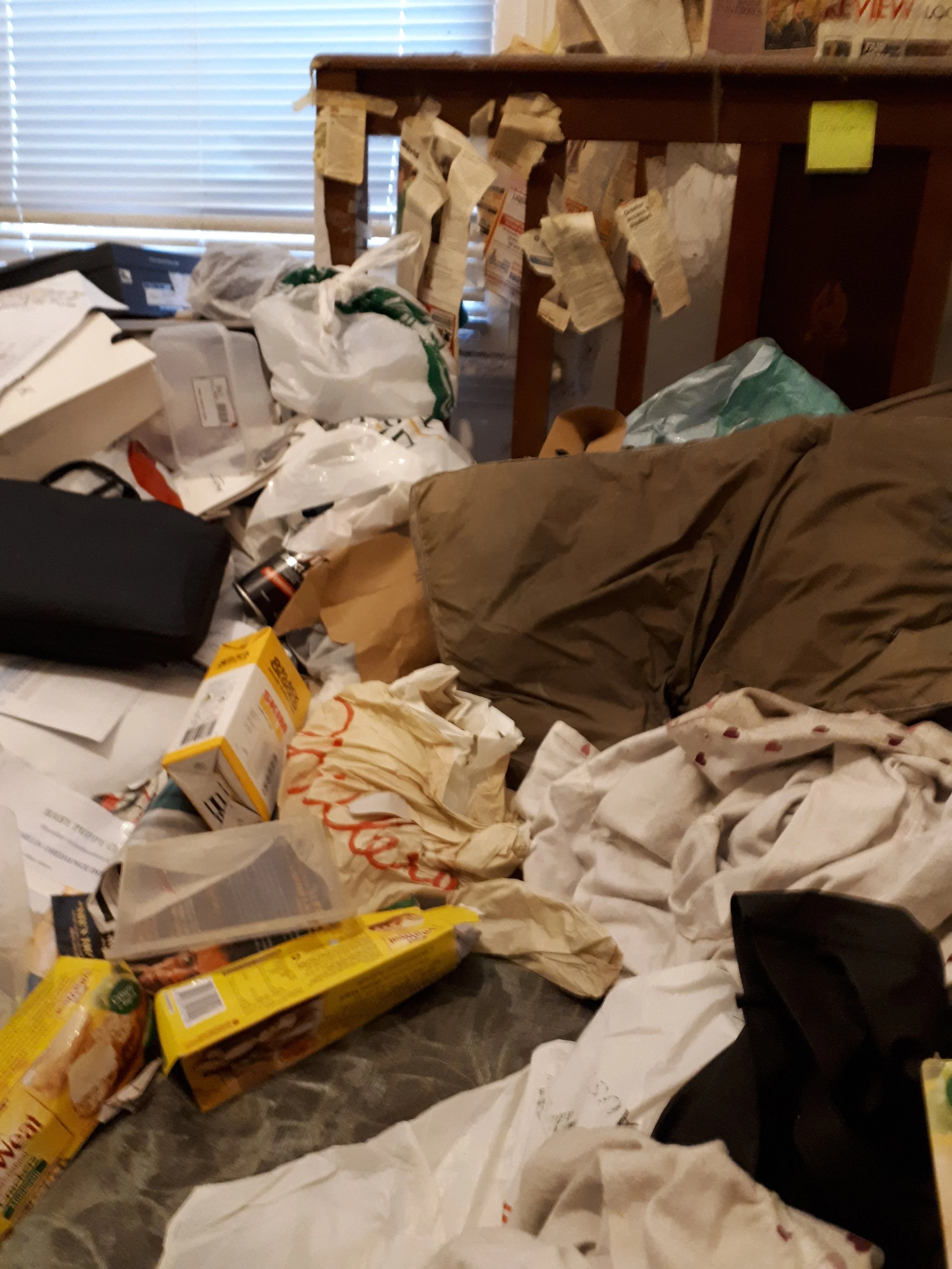 A cluttered room with papers, boxes, and packaging materials strewn across the floor and furniture, including a brown couch and a wooden table or shelf with notes attached.