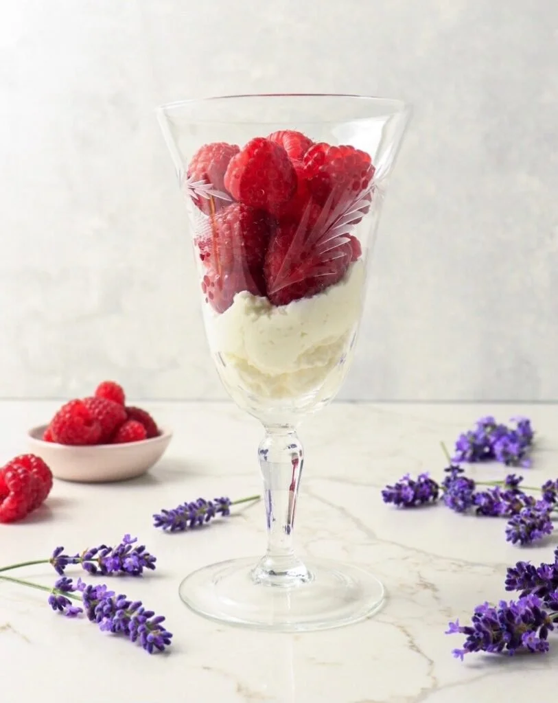 A glass dessert cup filled with fresh raspberries and a scoop of vanilla ice cream, with additional raspberries in a small bowl and sprigs of lavender on a white surface.