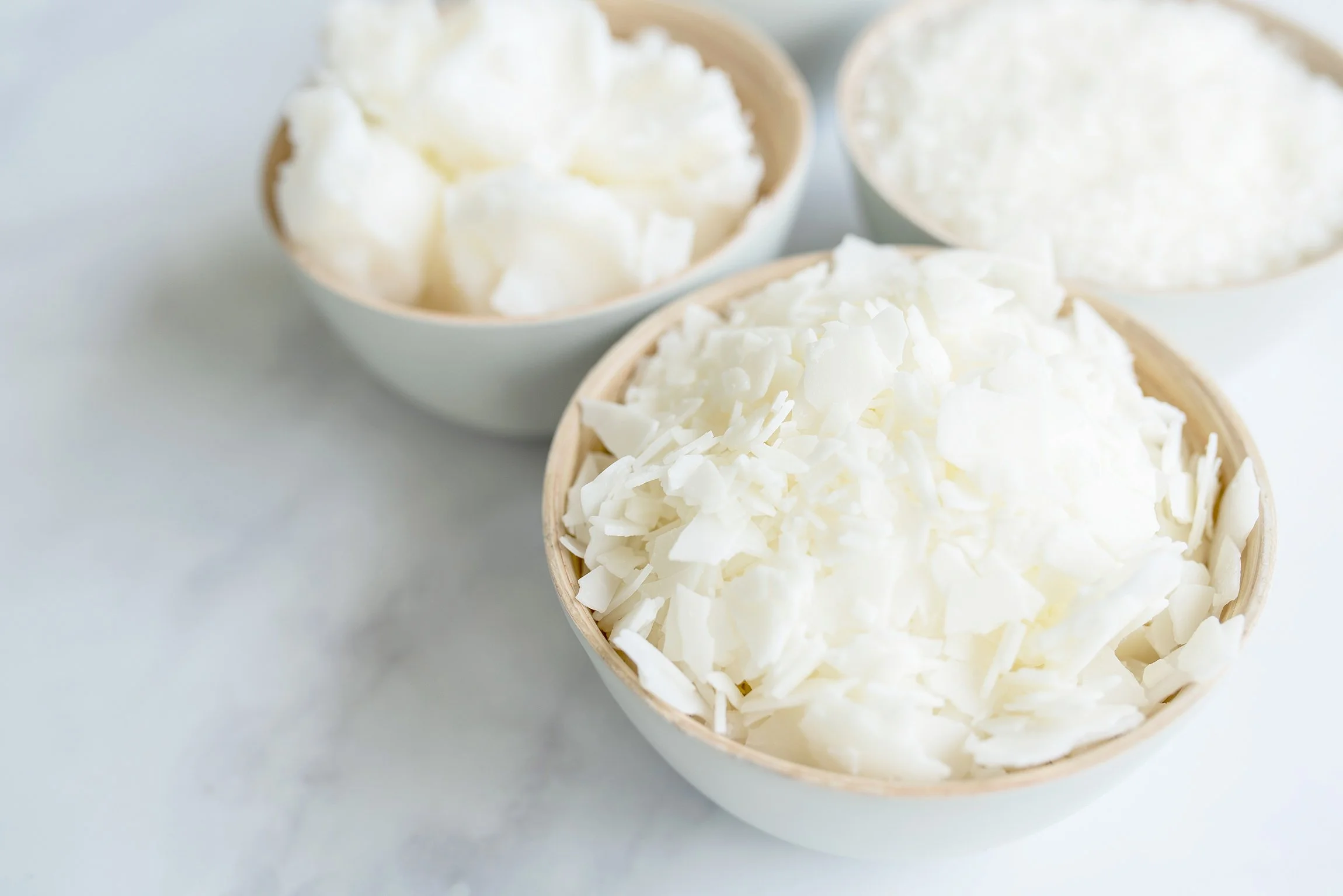 Close-up of three small bowls filled with flakes of soy wax, arranged on a light surface.