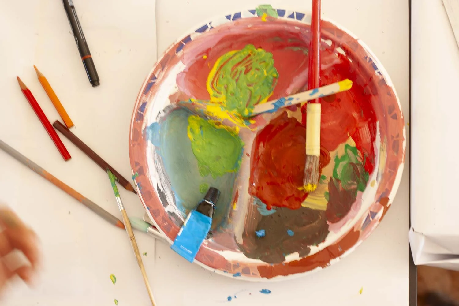 Une palette d'artiste avec des peintures de différentes couleurs et plusieurs pinceaux, certains enduits de peinture, sur une surface blanche, entourée de crayons de couleur.