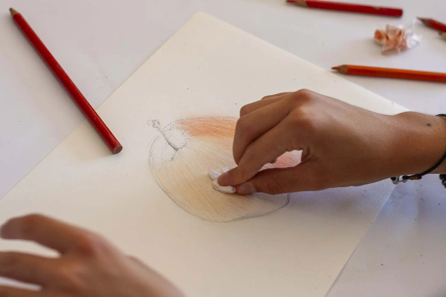 Une personne en train d'effacer un dessin d'un pomme colorée avec un tissu blanc sur une feuille blanche, entourée de crayons rouges.