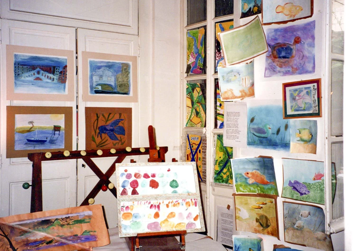 Une exposition d'œuvres d'art comprenant des peintures de paysages et de poissons, organisées sur des panneaux et un chevalet dans une pièce lumineuse.