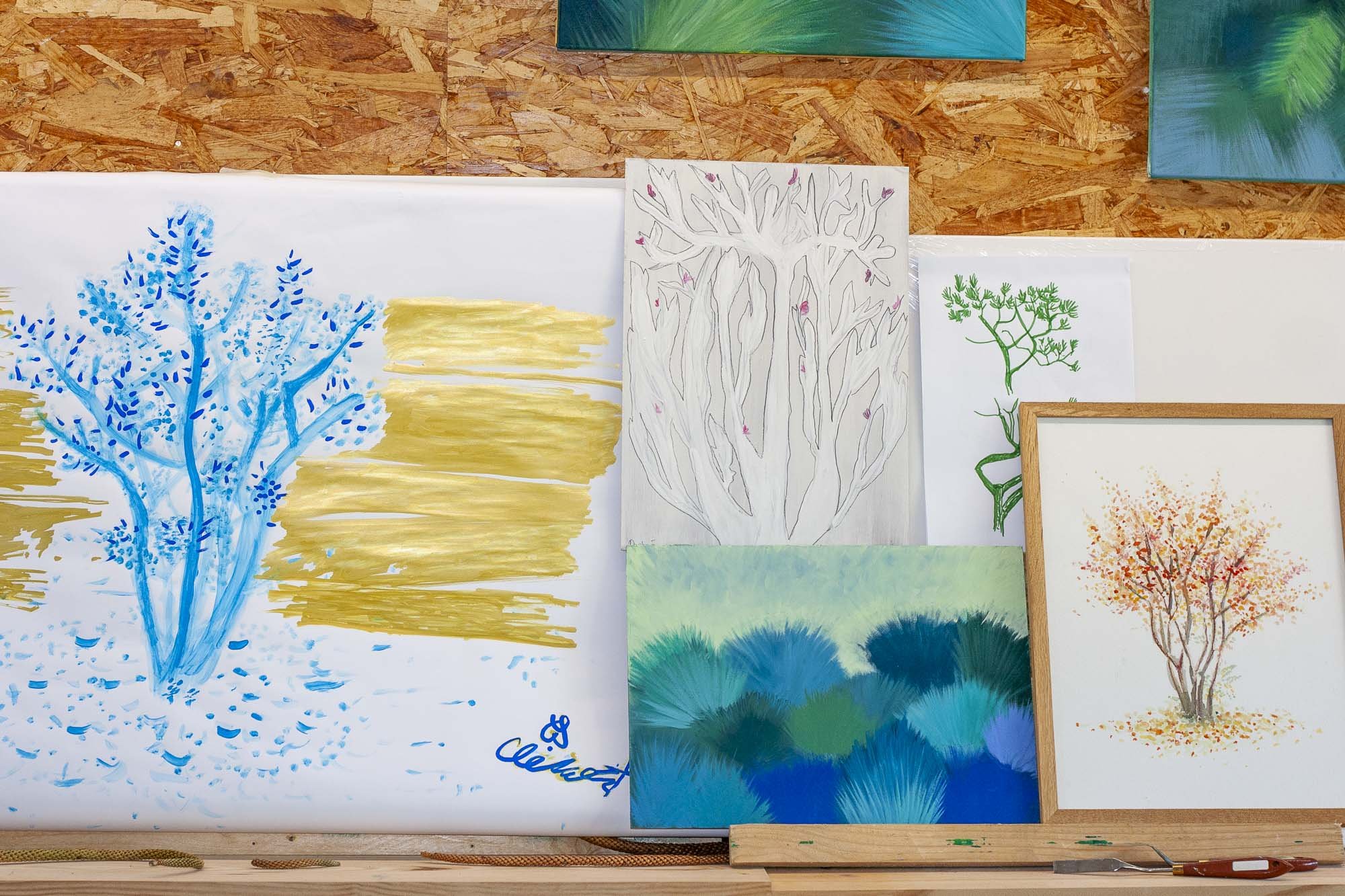 Dessins de paysages, notamment des arbres colorés, exposés sur un mur en bois au sein d'un atelier d'arts plastiques