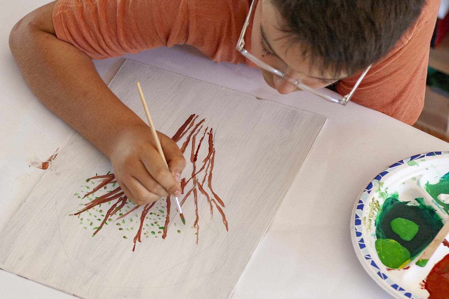 Un enfant en train de peindre une image de racines d'arbre sur un panneau en bois, utilisant un pinceau fin et des couleurs vertes et marron, avec un plateau de peinture à côté.