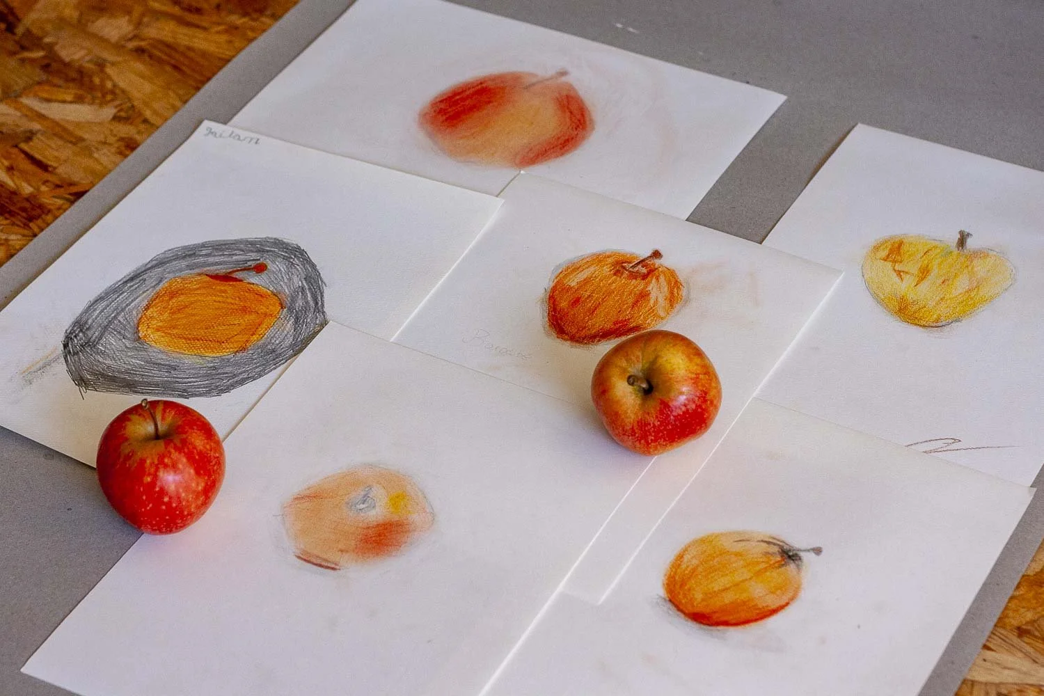 Dessins de pommes réalisés avec des crayons de couleur ou de peinture, accompagnés de vraies pommes, posées sur des feuilles de papier sur une table.