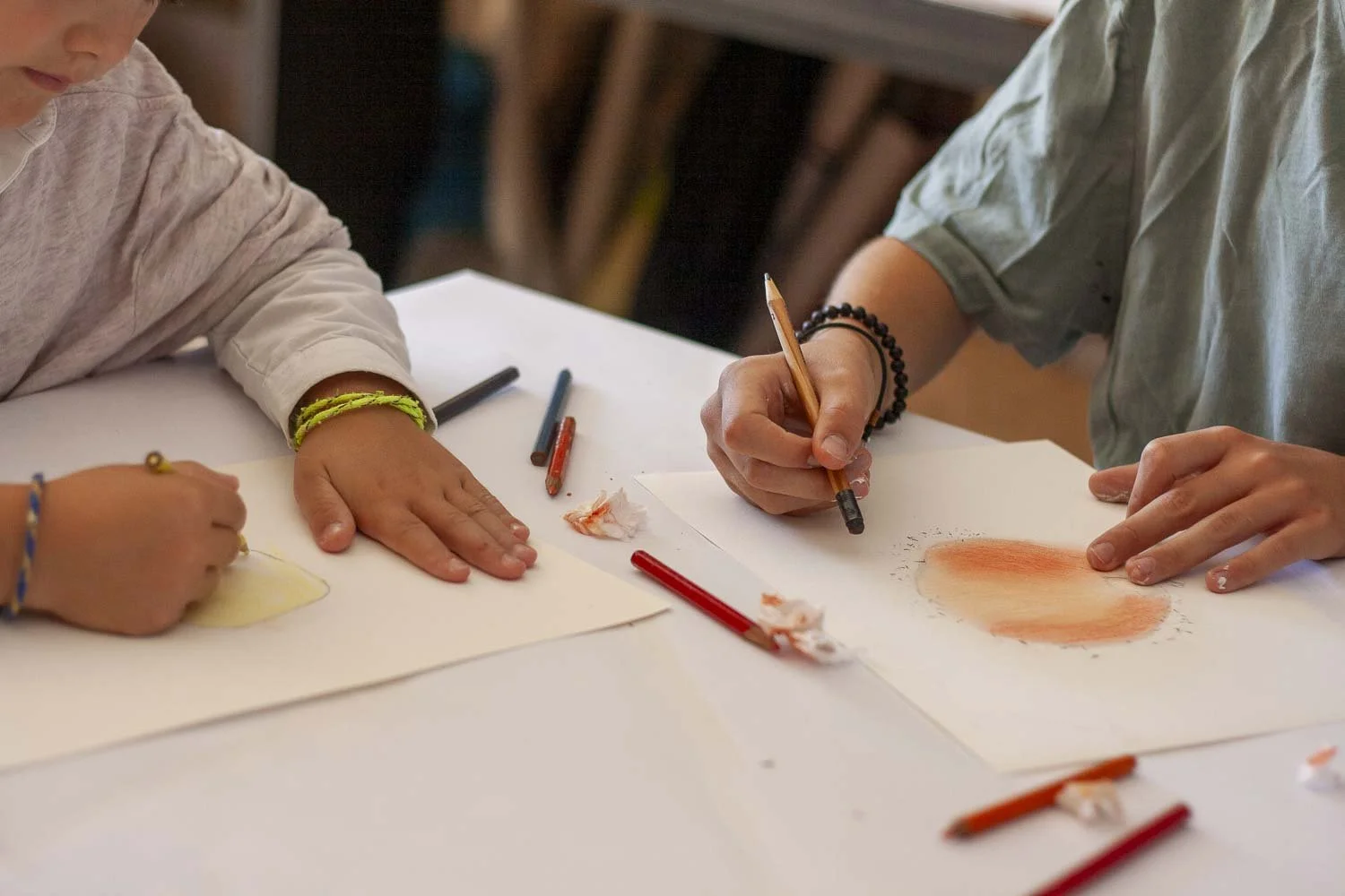 Deux enfants font du dessin avec des crayons de couleur sur des feuilles de papier blanc. Un dessin utilise des couleurs chaudes comme l'orange et le rouge.