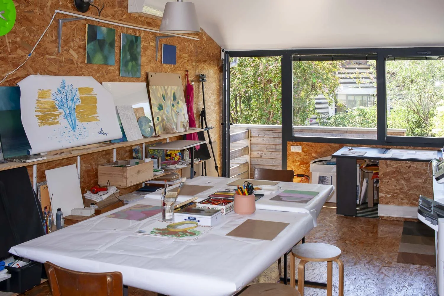 Atelier d'art avec table recouverte d'une nappe blanche, fournitures de peinture, échantillons, toiles, et grandes fenêtres laissant entrer la lumière naturelle, avec vue sur un jardin en arrière-plan.