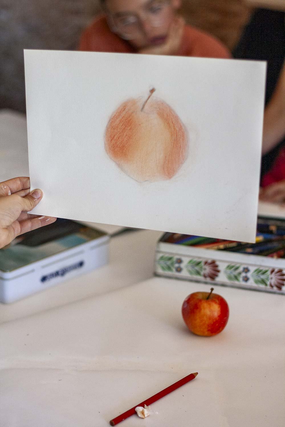 Une personne tient un dessin, réalisée à la craie ou à l'aquarelle, devant une vraie pêche posée sur une table. Un enfant regarde le dessin avec intérêt, derrière eux, on voit de cassettes ou boîtes de rangement, et un crayon rouge est posé sur la ta