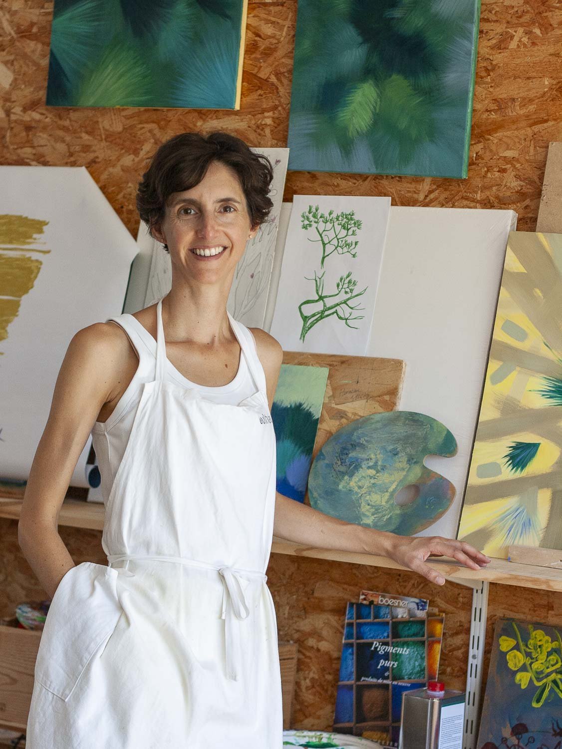 Une femme souriante portant un tablier blanc, debout dans un atelier d'art entourée de plusieurs peintures colorées.