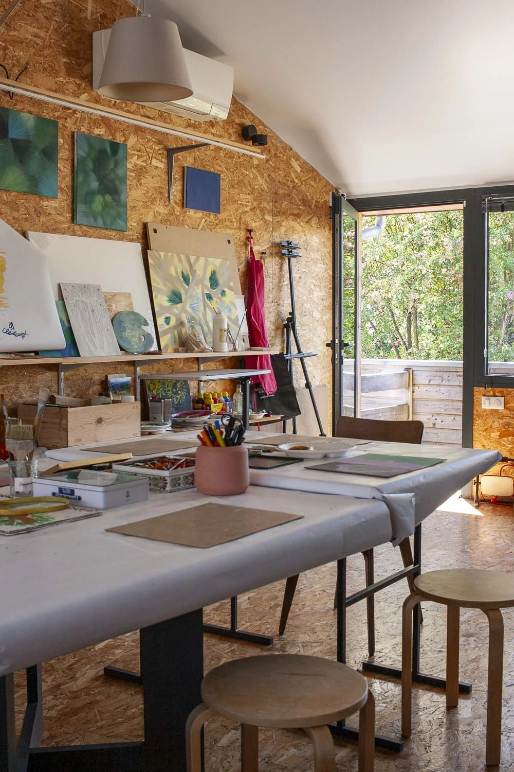 Atelier d'art avec un grand tableau blanc, des toiles, des pinceaux, et des fournitures artistiques sur une grande table, une baie vitrée laissant entrer la lumière naturelle, mur en bois aggloméré avec œuvres d'art