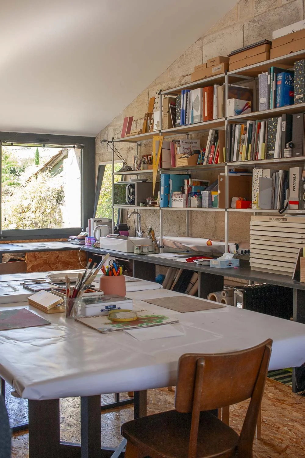 Un atelier ou espace de création avec une table recouverte de papier, des boîtes de crayons, des pinceaux, et des outils artistiques, avec une étagère pleine de livres et de fournitures, près d'une fenêtre qui laisse entrer la lumière naturelle.