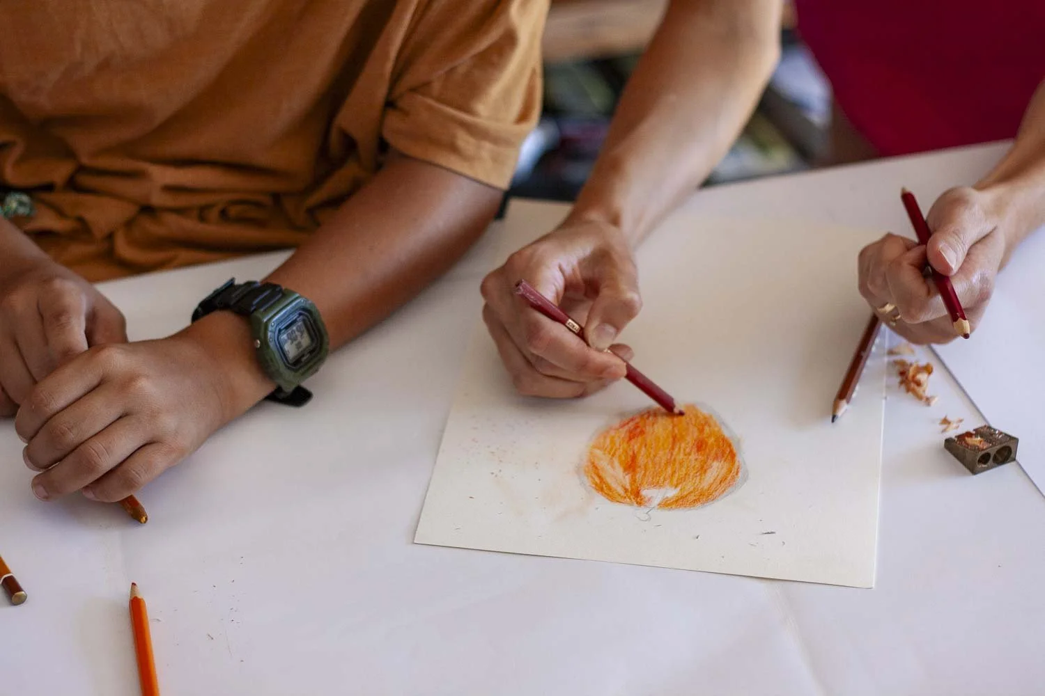 Deux personnes dessinent une pomme rouge orangée avec des crayons rouges sur une feuille blanche.