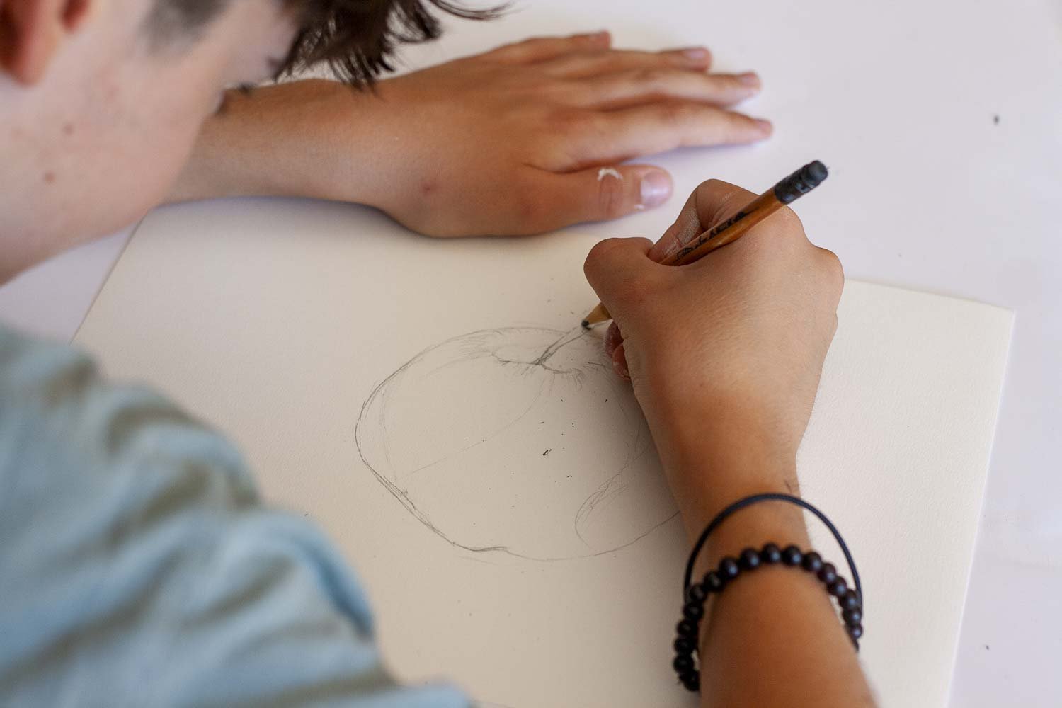 Un enfant dessine une pomme sur une feuille blanche, en utilisant un crayon, le bras repose sur la table.