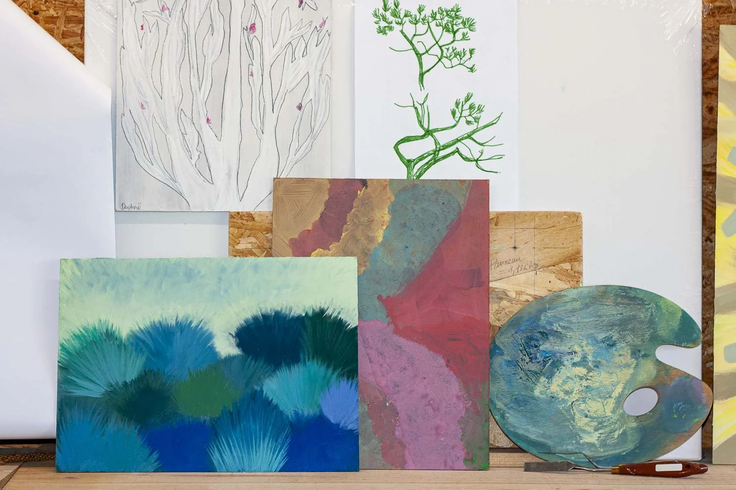 A collection d'œuvres d'art comprenant des dessins, des peintures et une palette de peinture, disposées sur un tableau en bois et un fond blanc.