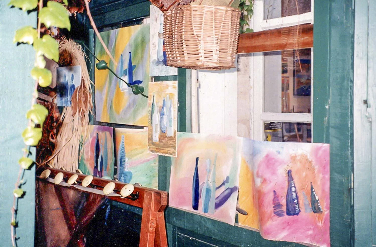 Une galerie d'art avec des peintures abstraites colorées exposées sur un mur vert. Un panier en osier est suspendu, et il y a des guirlandes lumineuses décoratives autour.