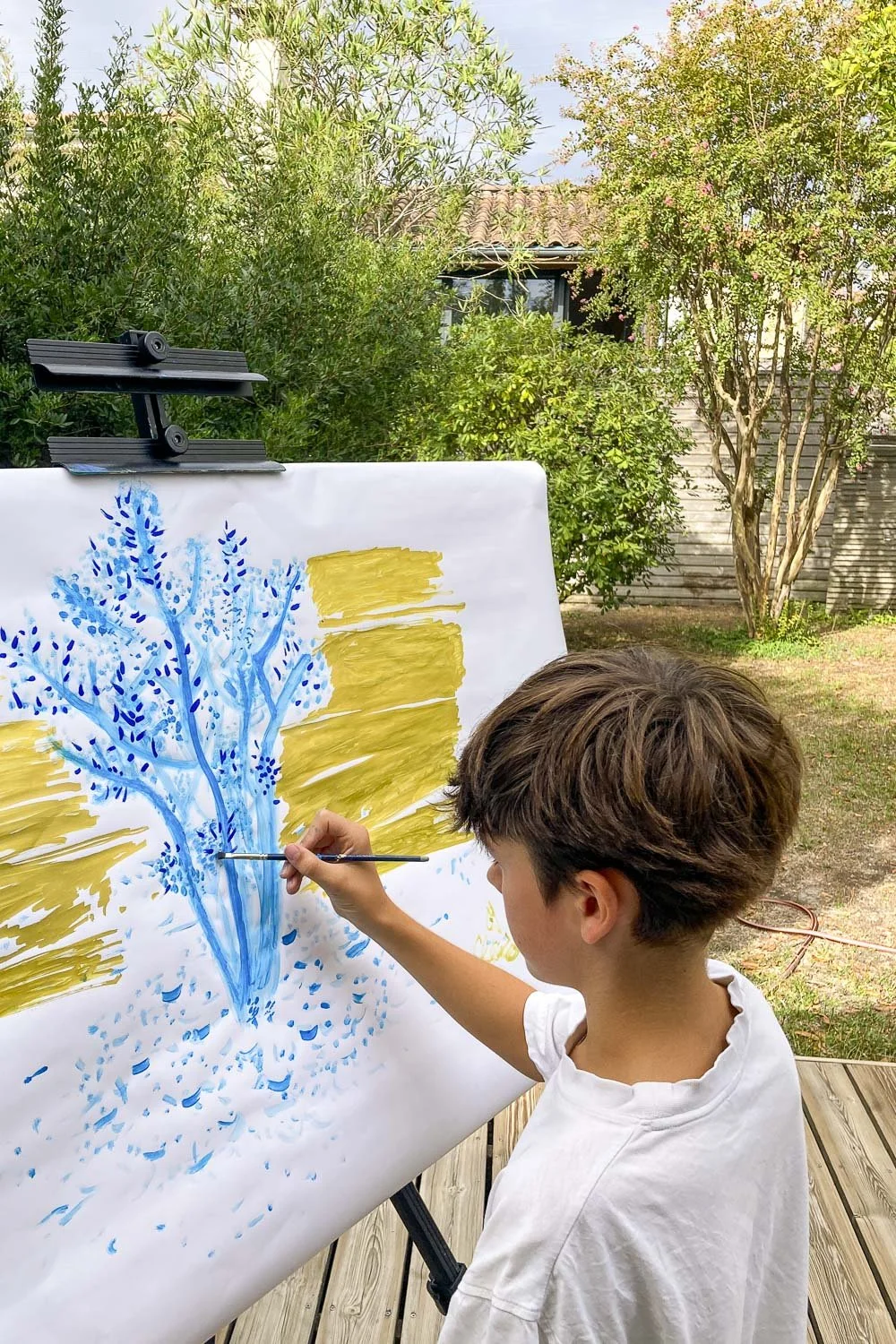 Un garçon peint un arbre bleu sur une toile à l'extérieur, avec un fond de jardin et un ciel nuageux.