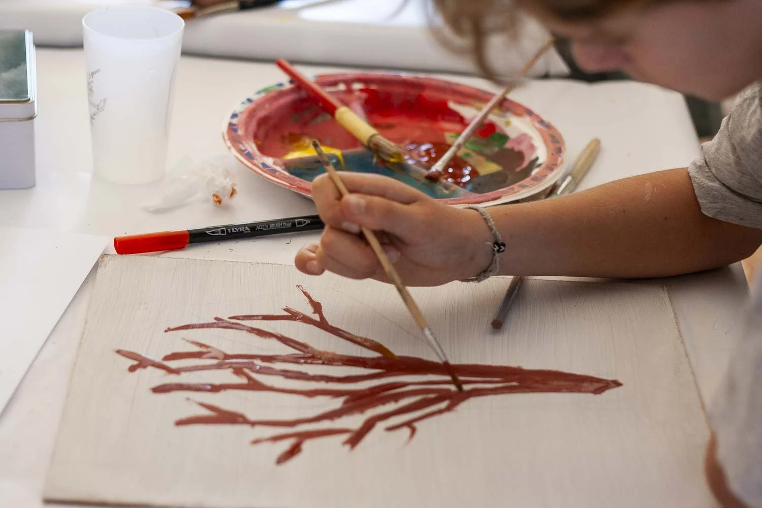 Une personne en train de peindre un arbre nu avec des branches rouges sur une toile blanche. Divers pinceaux et une palette de peinture avec plusieurs couleurs sont visibles sur la table.