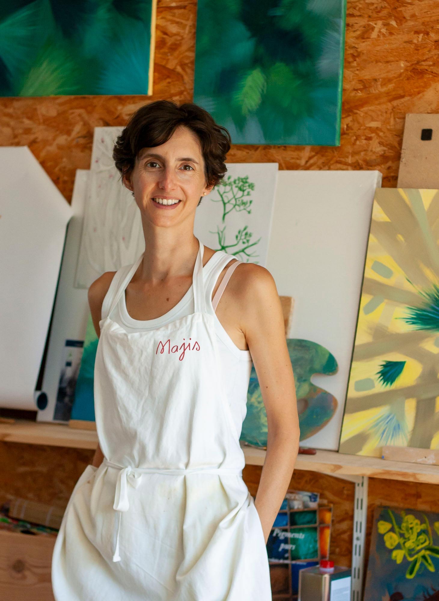 Une femme souriante portant un tablier blanc dans un atelier d'art avec des peintures colorées en fond.