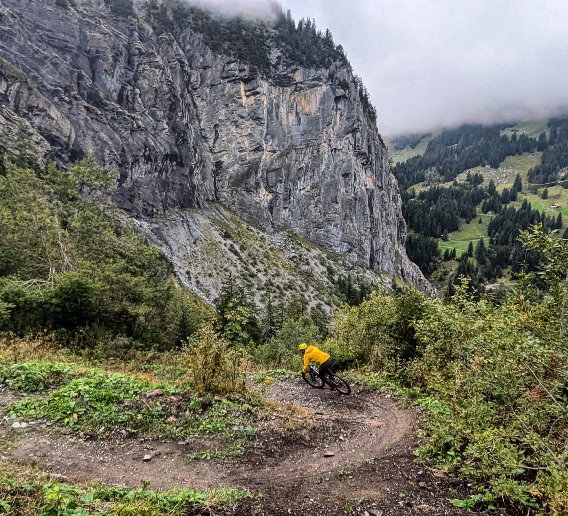 A person wearing a yellow jacket and a helmet riding a mountain bike down a dirt trail in a forested area, with tall rock cliffs and green hills in the background, and low-hanging clouds or mist in the sky.