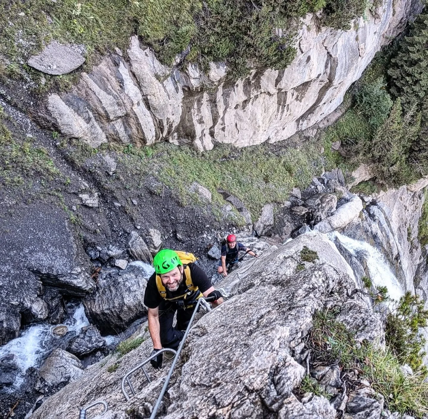Two people rock climbing on a steep rocky waterfall, wearing helmets and climbing gear, climbing up a mountain terrain with water flowing below.