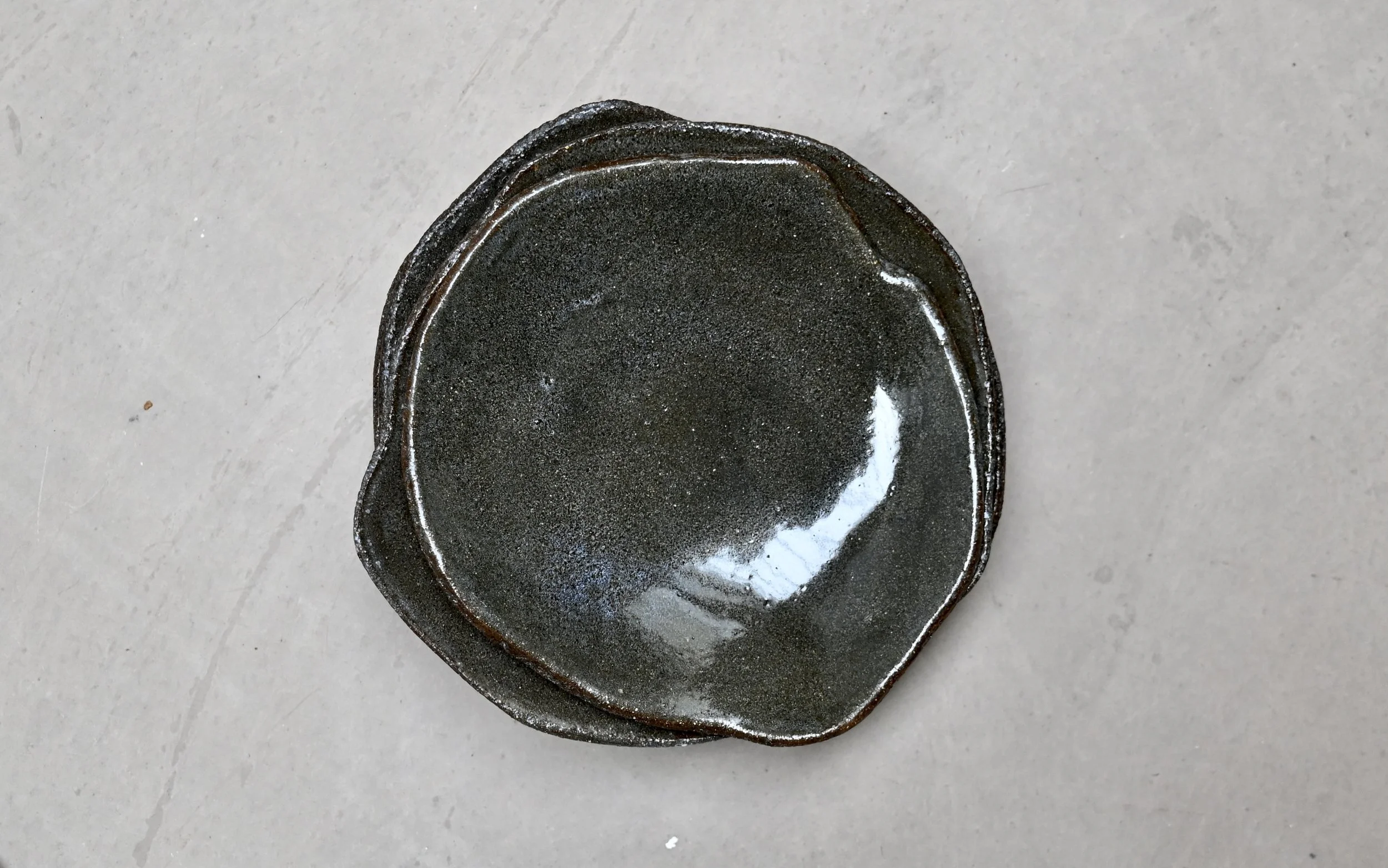 Assiettes originales, réalisées par Jeanne Yaa Stenmanns en grès rouge et émaillées en vert. Elle sont empilées sur une surface en béton clair.