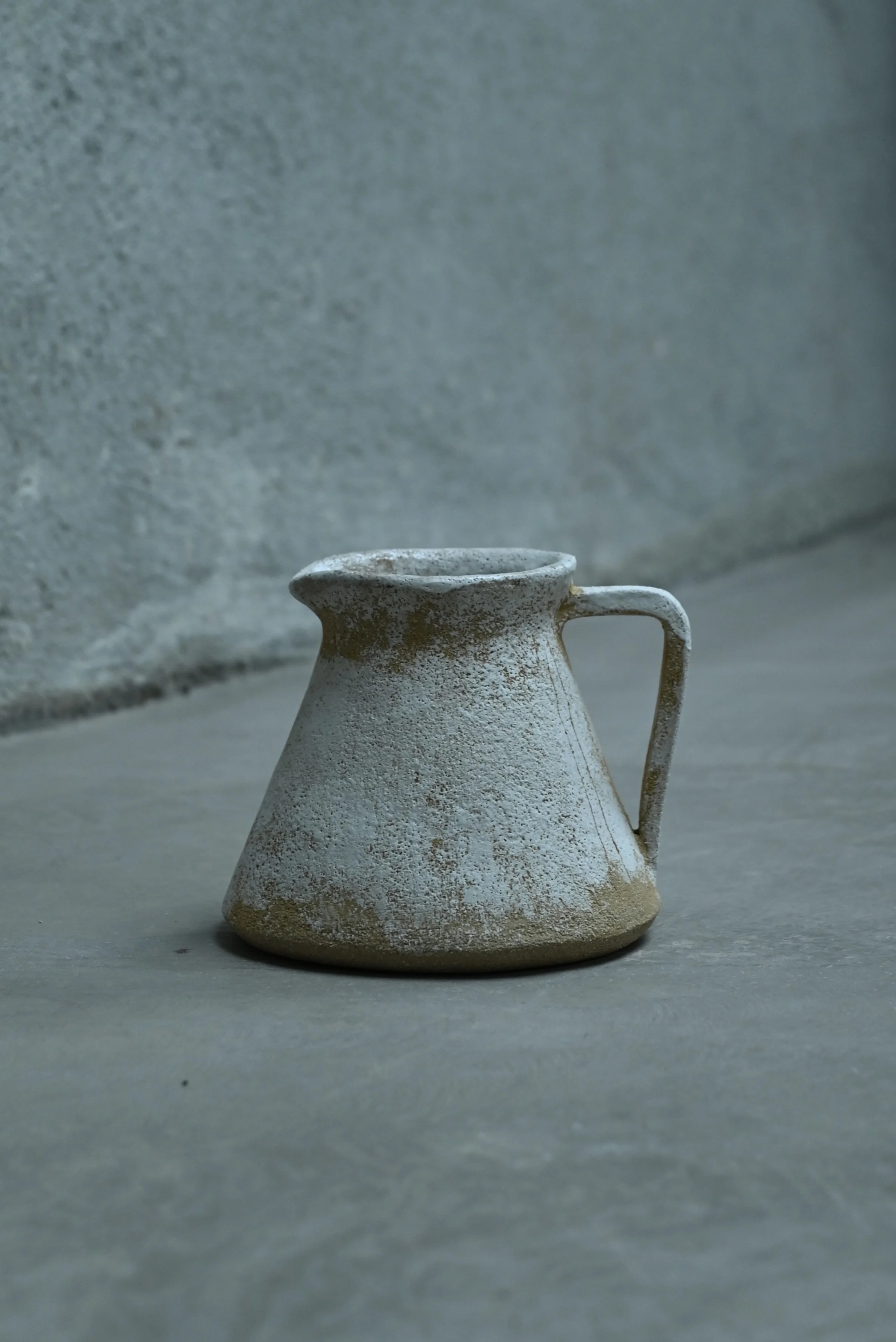 Pichet en grès roux réalisé au modelage par Jeanne Yaa Stenmanns, placé sur une surface en pierre, contre un mur en béton gris, dans une ambiance minimaliste et brute. Le pichet a été décoré à l'engobe blanche. 