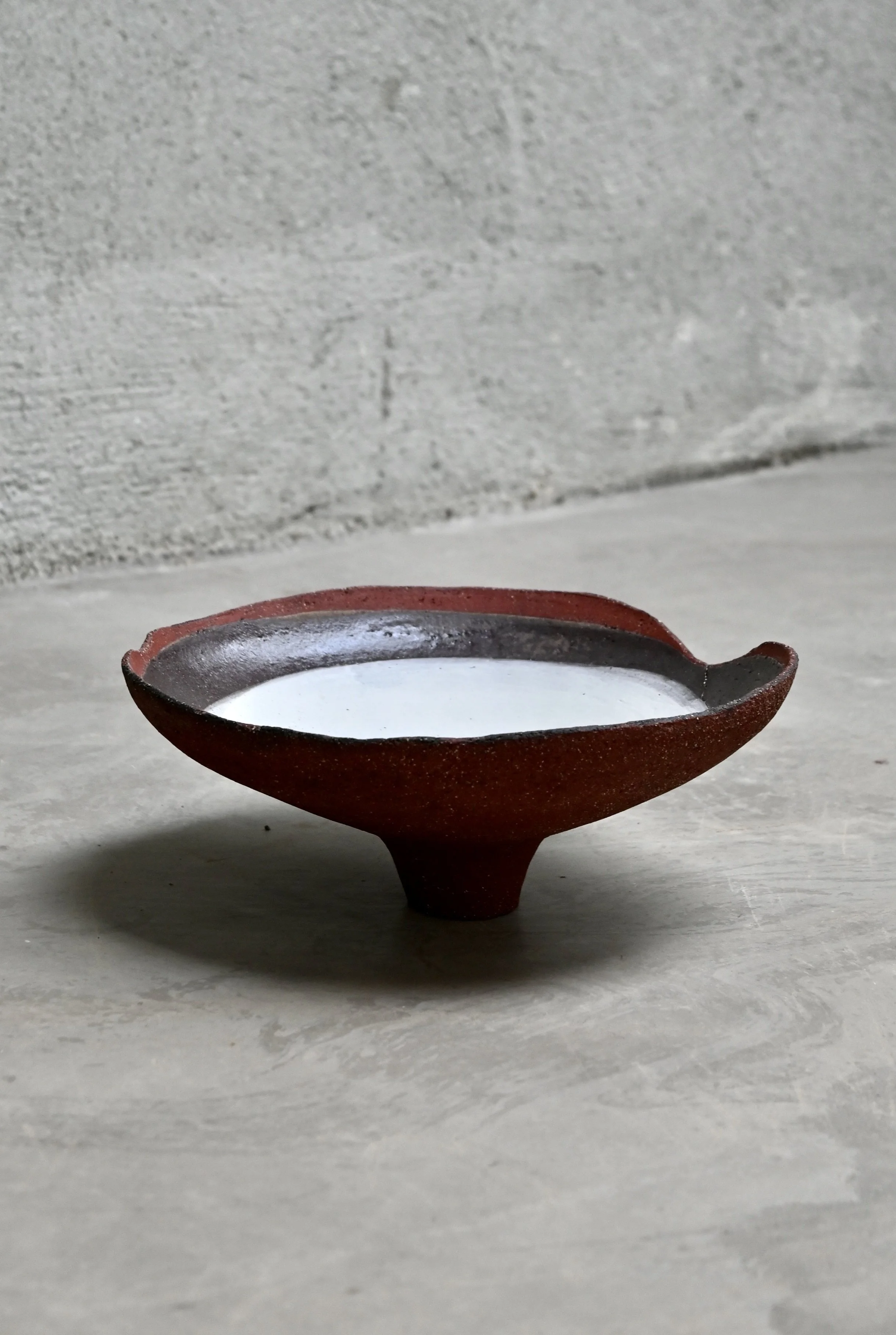 Vasque en grès rouge, décor à l'engobe blanche, réalisée au modelage par Jeanne Yaa Stenmanns.  La céramique est placée sur une surface en pierre ou en béton, avec un fond en mur gris.