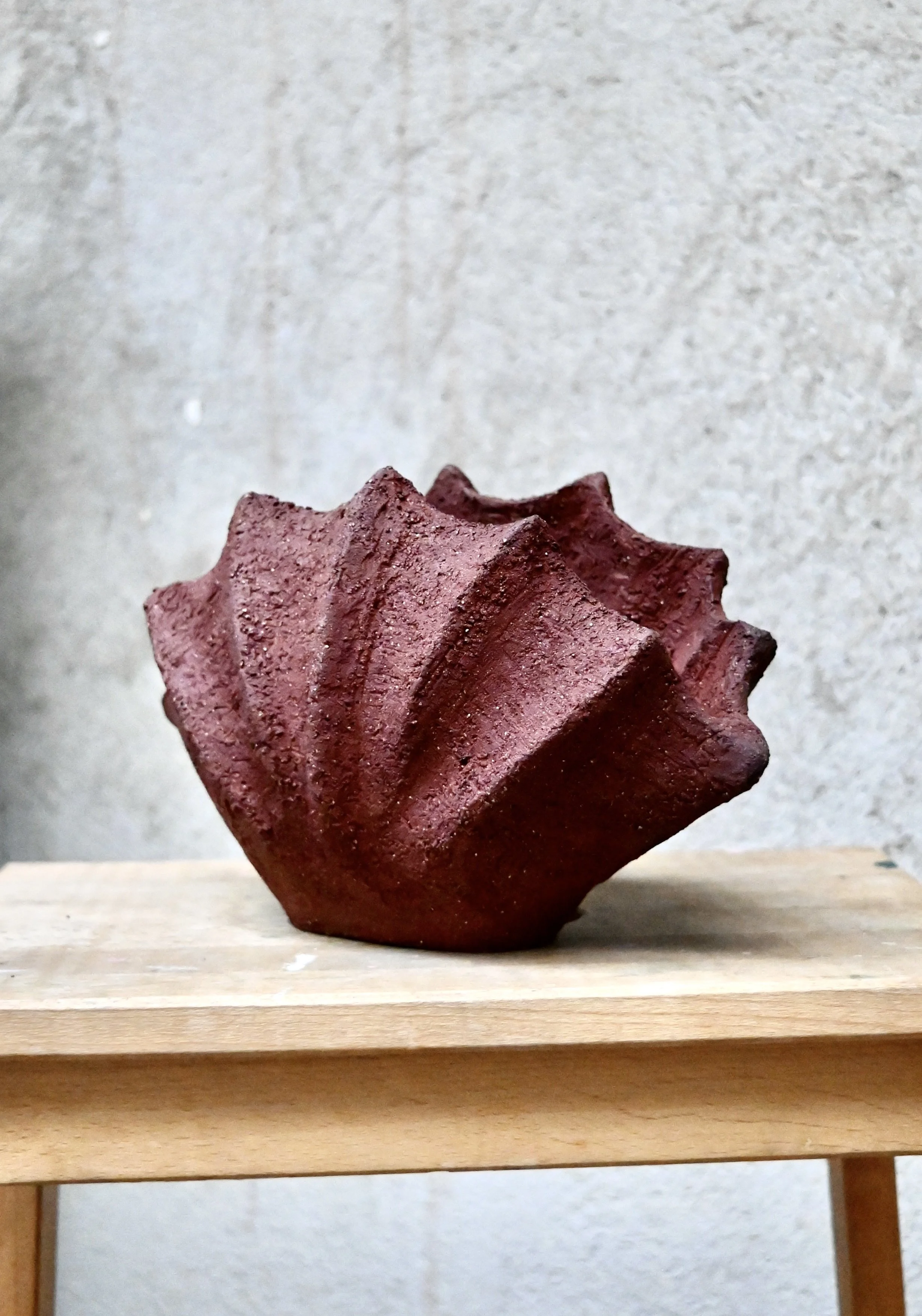 Une sculpture en forme de coquillage en grès rouge, posé sur une table en bois. Pièce sculpturale réalisée par Jeanne Yaa Stenmanns au modelage. 