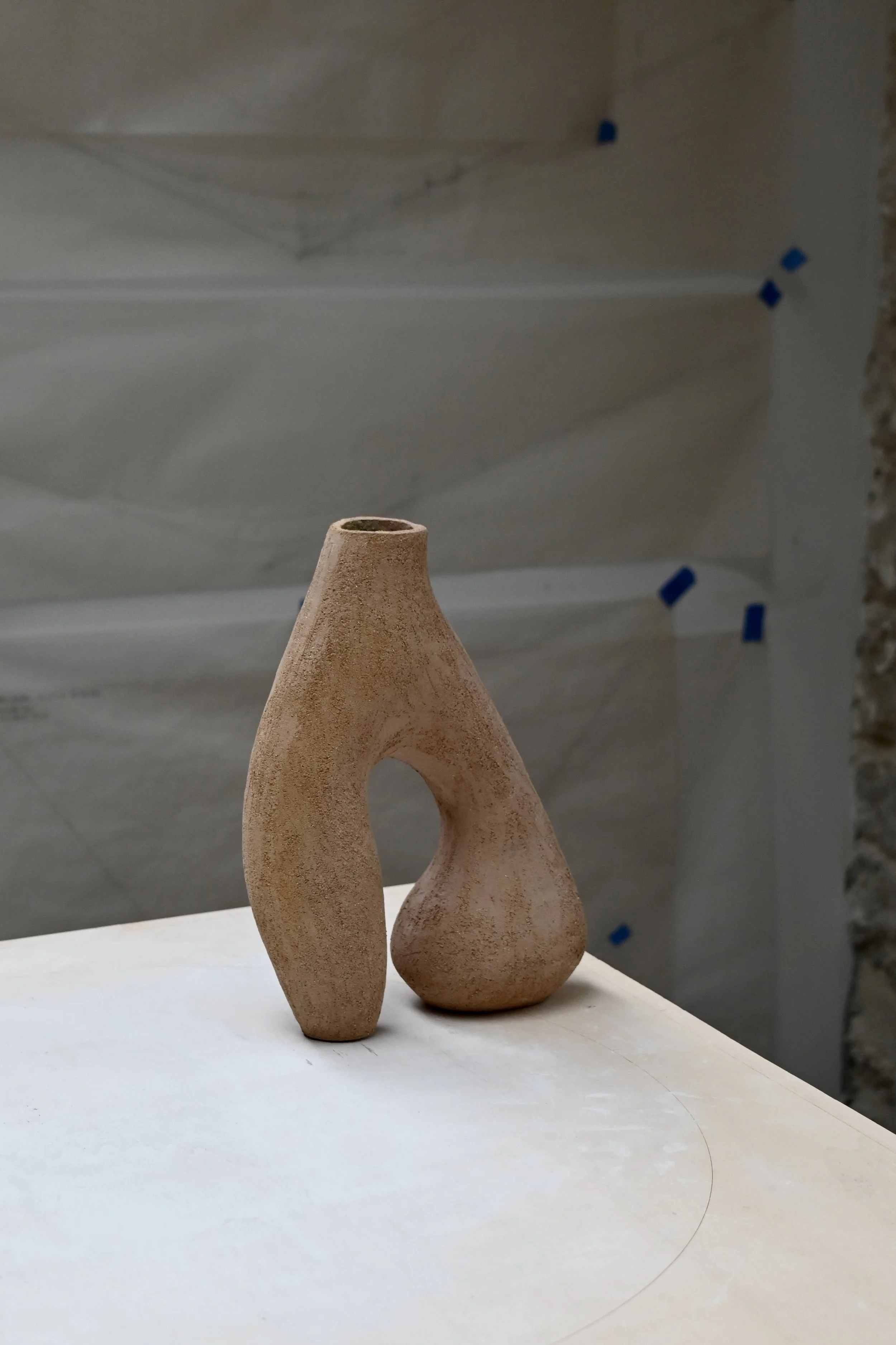 Vase en grès roux avec une forme fuselée et un trou circulaire à son extrémité, placée sur une surface blanche, avec un mur en pierre apparent en arrière-plan. Forme inspirée de Valentine SCHLEGEL, réalisée par Jeanne Yaa Stenmanns.