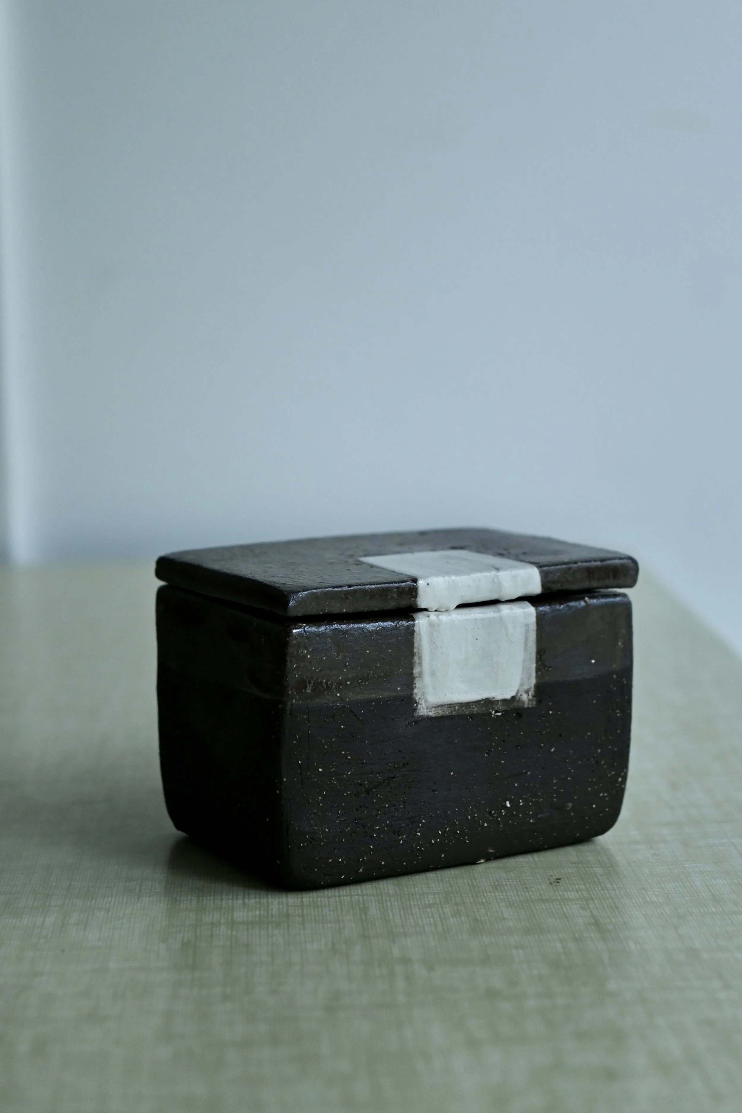 Une boîte en grès noir avec un couvercle noir et une bande blanche, posée sur une surface verte. Le fond est gris clair.