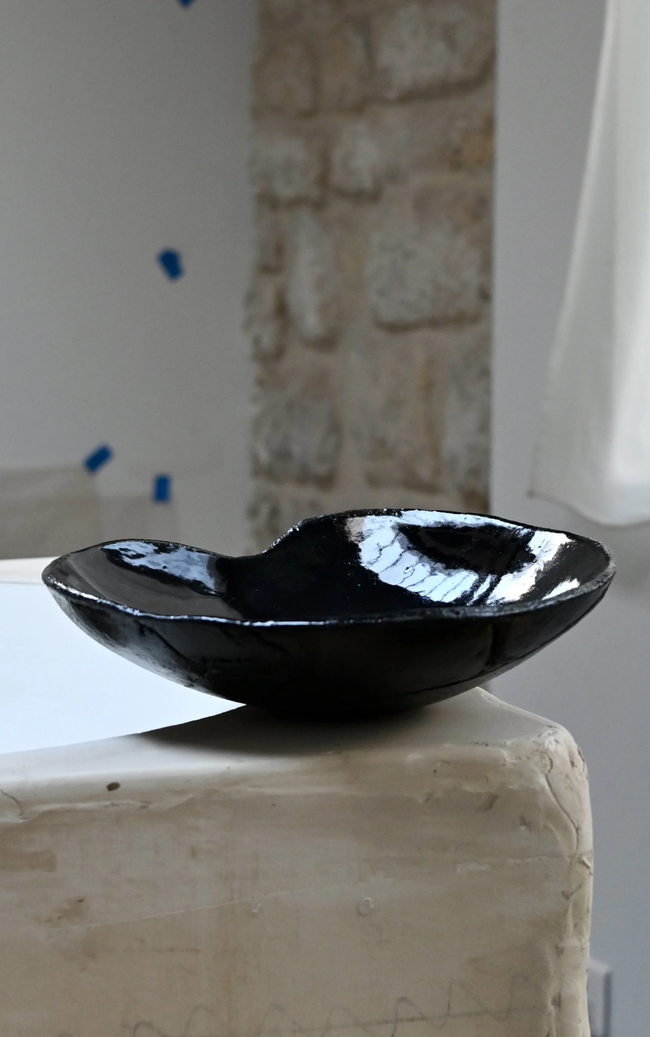 Plat en céramique avec une surface brillante, posée sur une base en pierre ou en céramique beige, devant un mur en pierre rustique et une porte blanche. Pièce réalisée par Jeanne Yaa Stenmanns au modelage. 