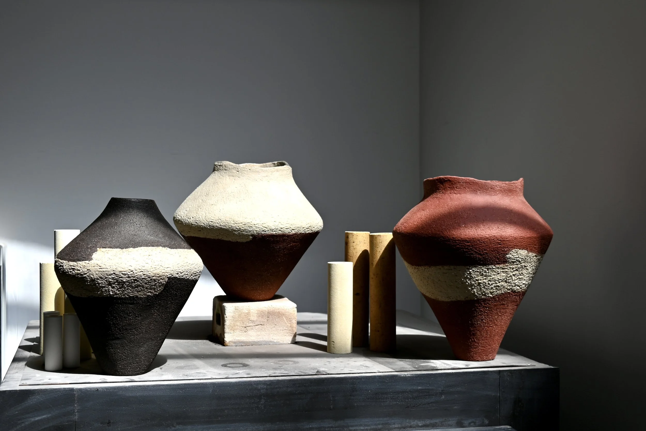 Vases en grès rouge, noir et blanc réalisés au modelage par Jeanne Yaa Stenmanns. Les vases en céramique sont de différentes tailles et couleurs, disposés sur une surface avec des supports cylindriques.