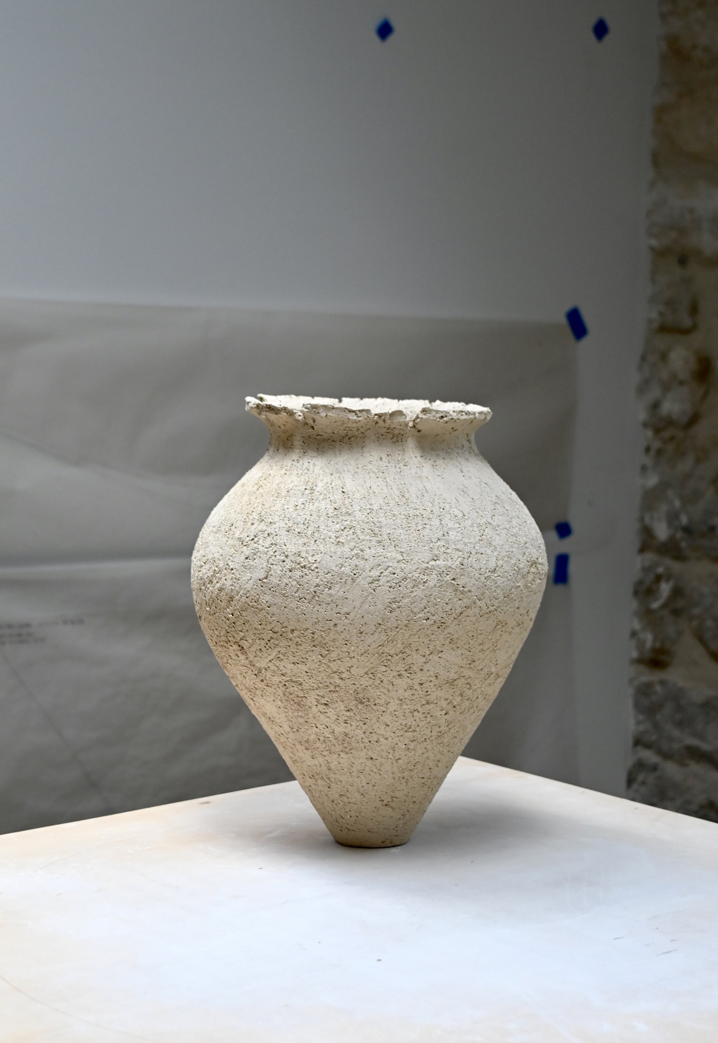 Très beau vase en grès blanc chamotté, crée par Jeanne Yaa Stenmanns au modelage, présenté sur un fond sombre avec certains éléments en pierre visible à droite. Aspect brut et rugueux. 
