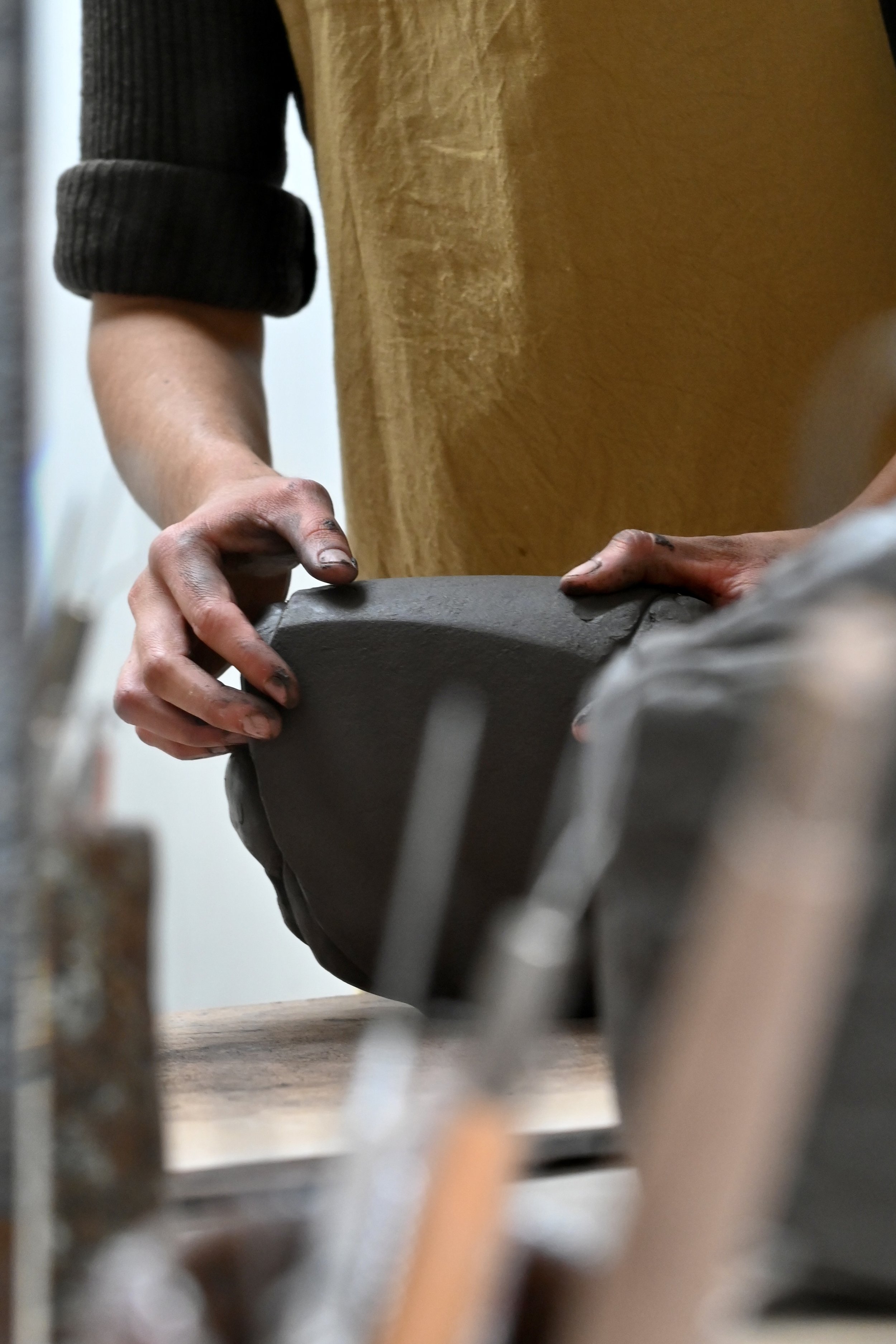Belle image des mains de Jeanne Yaa Stenmanns entrain de pétrir sa terre (un grès noir), sur sa table de travail au sein de l'atelier. On y aperçoit quelques outils.
