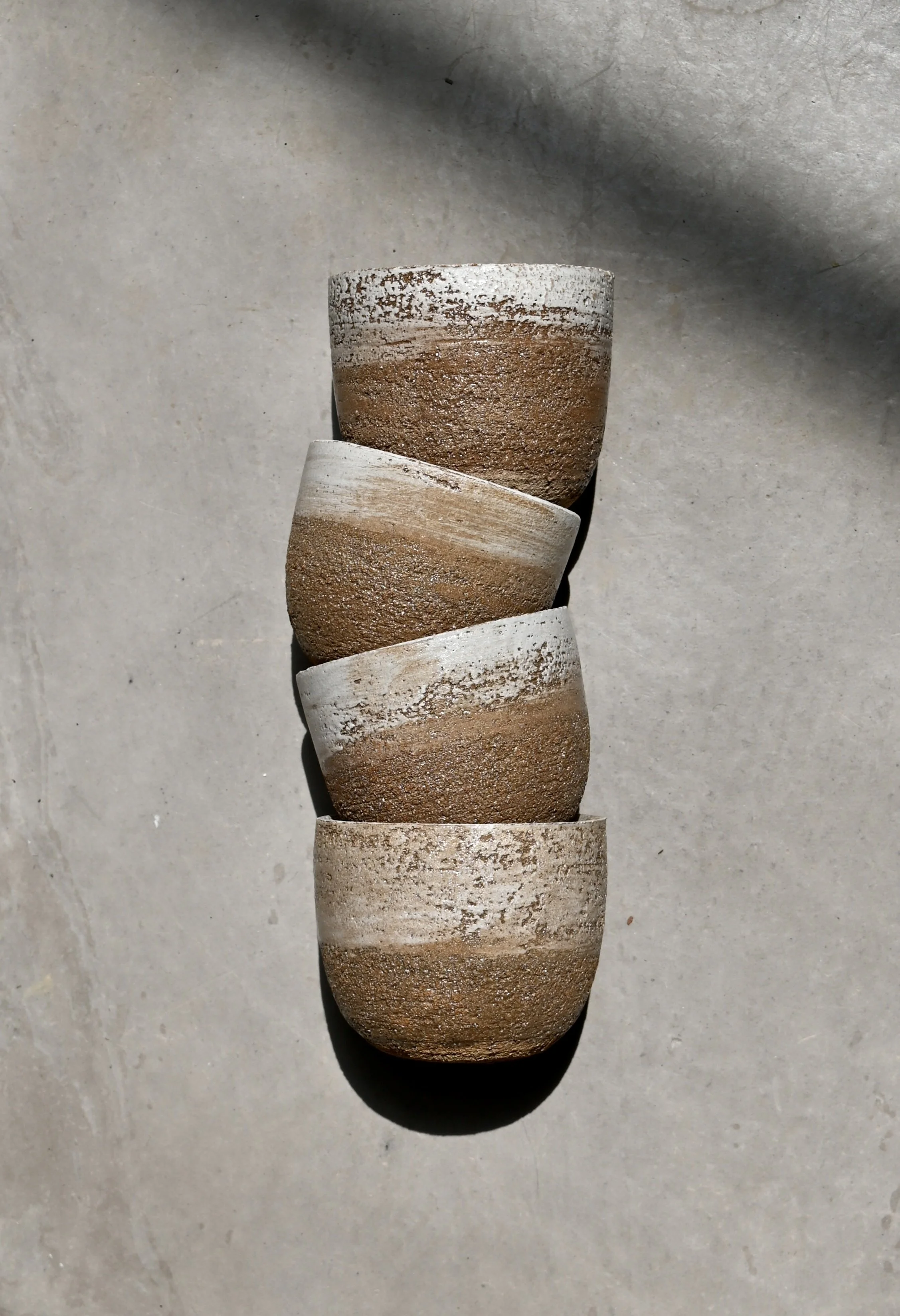 Une rangée de six tasses en céramique, chacune avec une finition rustique et une couleur variée, allant du blanc cassé au brun, placée sur un sol en béton. Réalisées au tournage par Jeanne Yaa Stenmanns. 
