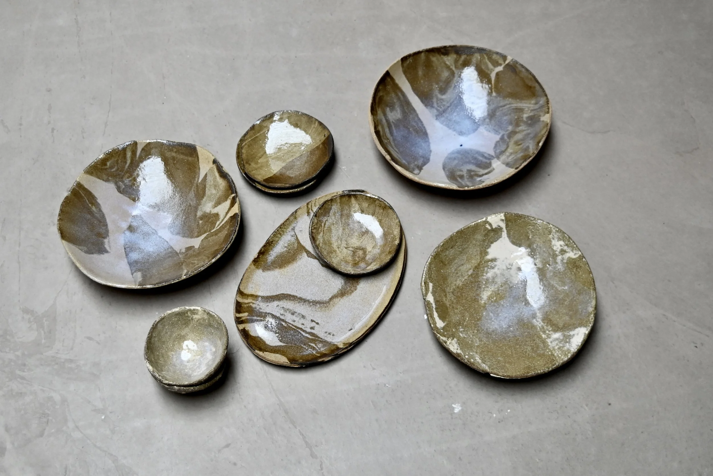 Une collection de petites assiettes en céramique avec un motif marbré de couleurs marron, beige et blanche, posées sur une surface grise. Assiettes et plats réalisés au modelage par Jeanne Yaa Stenmanns. 