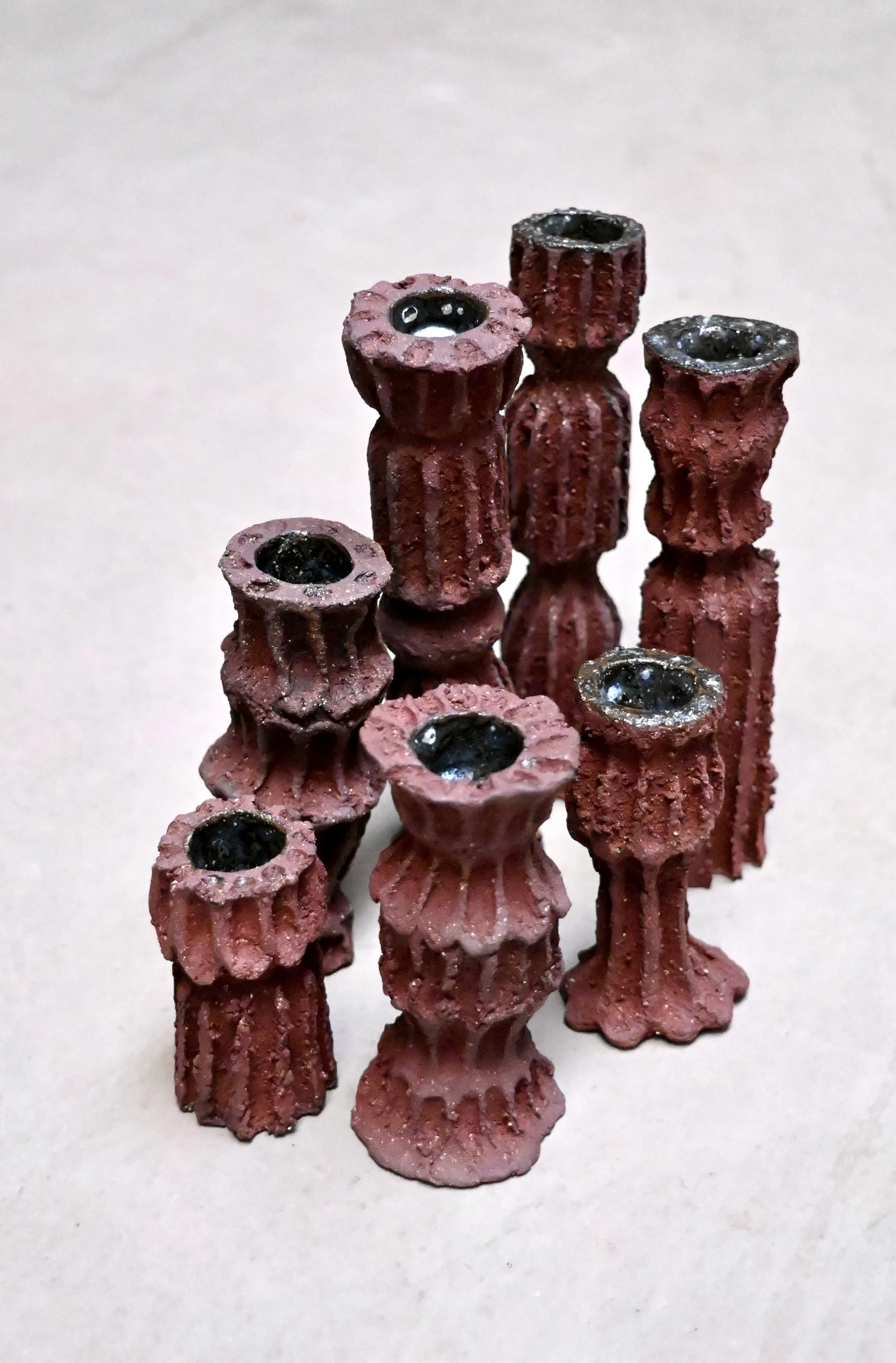Groupe de petits objets en forme de colonnes, de couleur rouge, avec une surface textured. Chacun a un trou en haut, certains avec un centre noir brillant, ressemblant à de petites vases ou tiges en miniature.