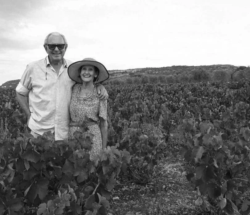 An elderly man and woman smiling in a vineyard, standing among grapevines, with the woman wearing a wide-brimmed hat.