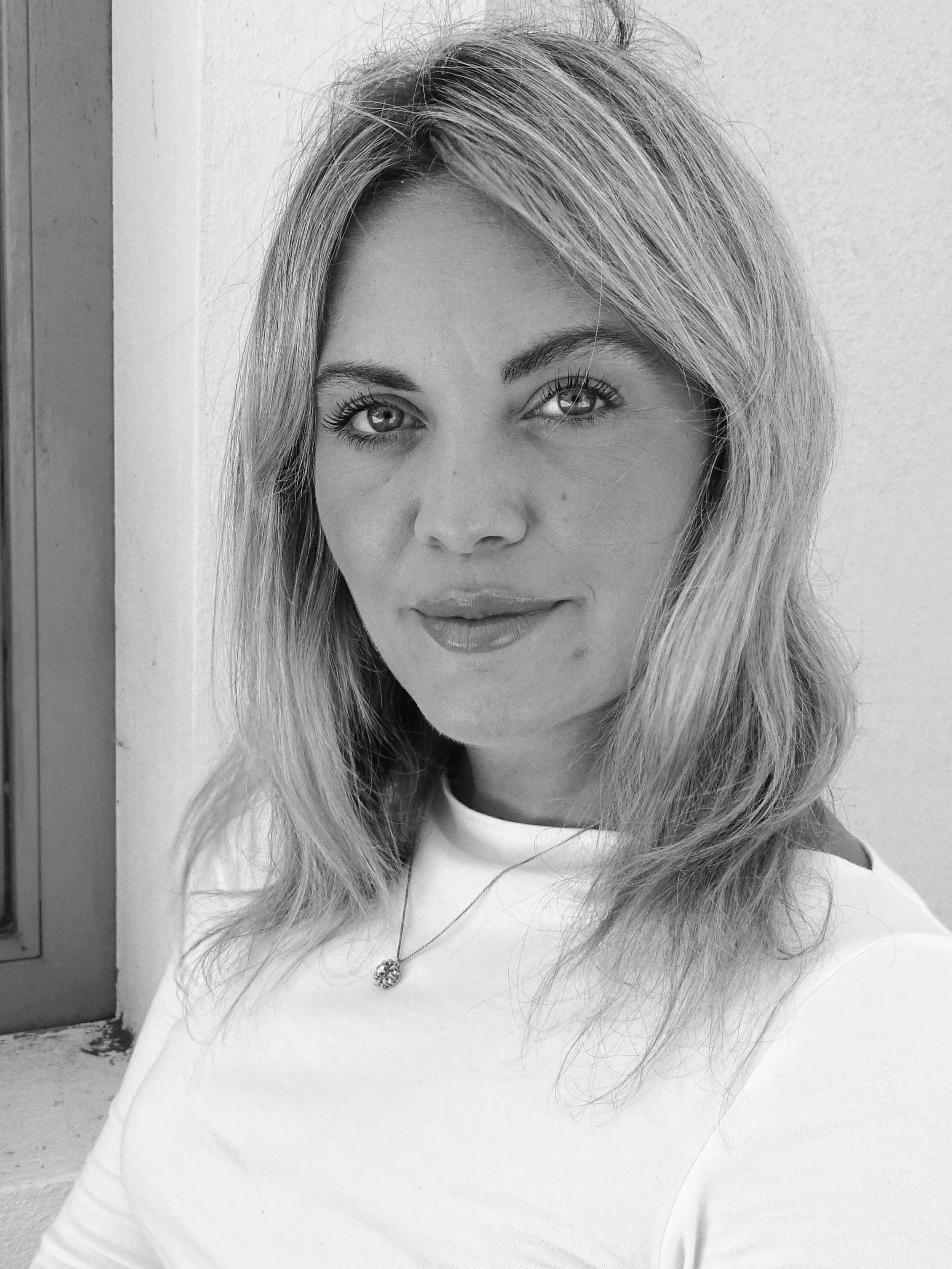 A black and white photo of a woman with long, wavy hair, wearing a light-colored top, standing outdoors near a wall.