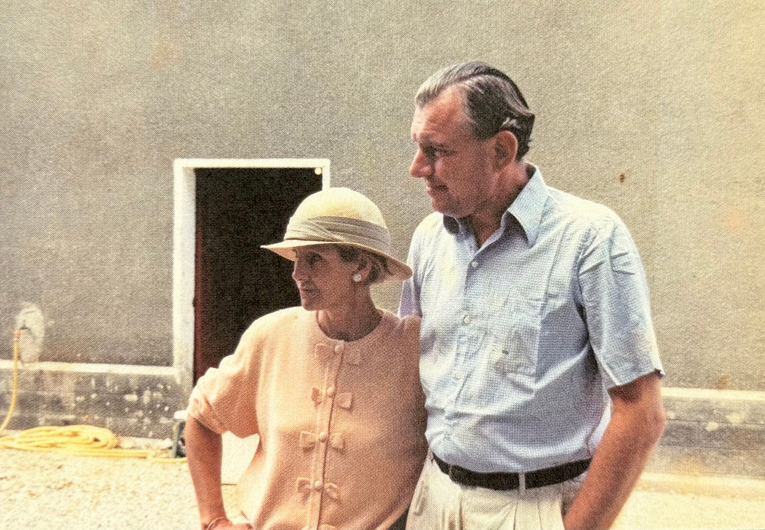 A woman wearing a white hat and a man with dark hair and a mustache, both dressed in light-colored clothing, are standing outdoors near a grill with food. The woman is looking at the man, who is looking slightly to the left. There is a window and some outdoor furniture or objects in the background.
