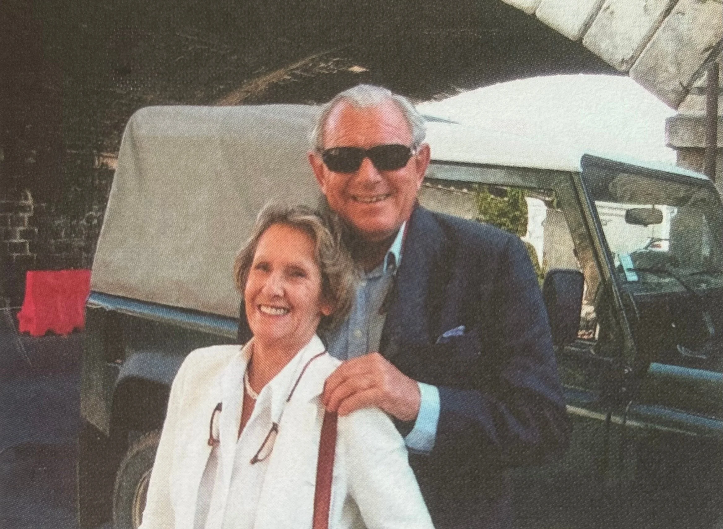 A man wearing sunglasses and a suit, smiling, standing next to a woman with short curly hair, smiling, and holding his jacket in a casual outdoor setting.